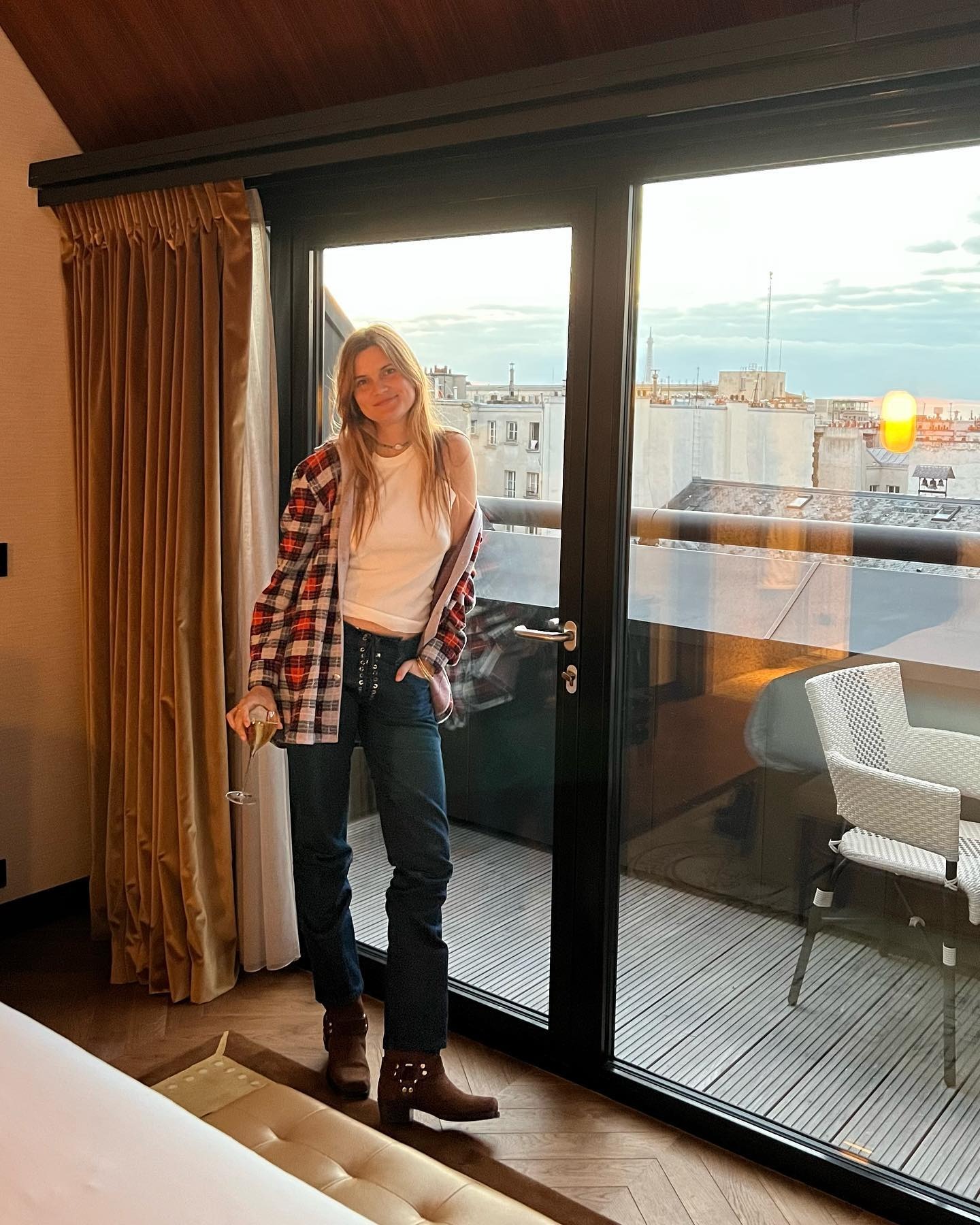 French writer, editor, and creative Eugenie Trochu poses in a penthouse space in Paris wearing a plaid button-down shirt, white T-shirt, lace-front jeans, and brown suede moto boots