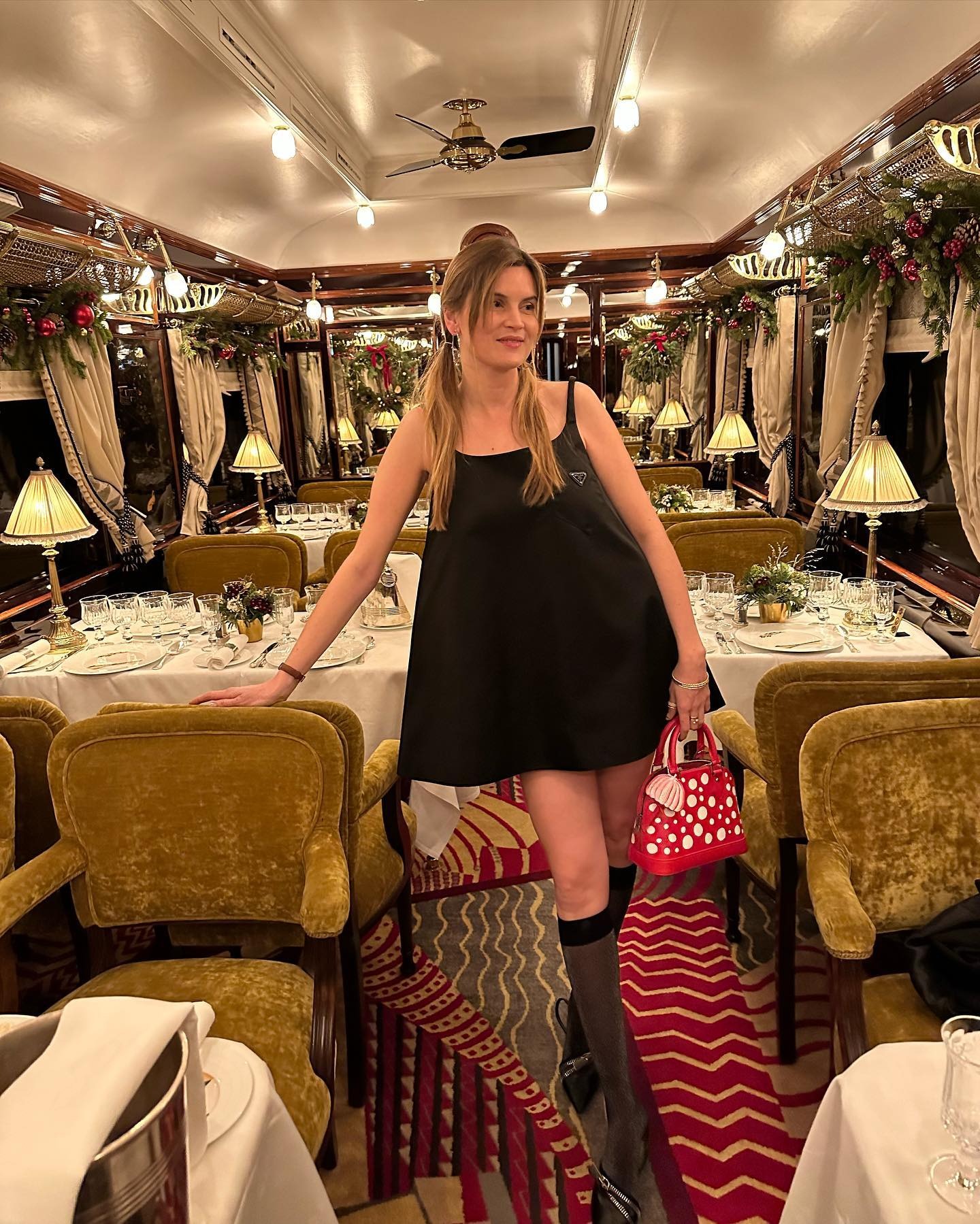 writer, editor, and fashion creative Euge&amp;#769;nie Trochu poses on the Christmas Orient Express Paris/Courchevel wearing a black Para mini dress, a white and red Louis Vuitton x Yayoi Kusama Alma bag, knee-high socks and heels