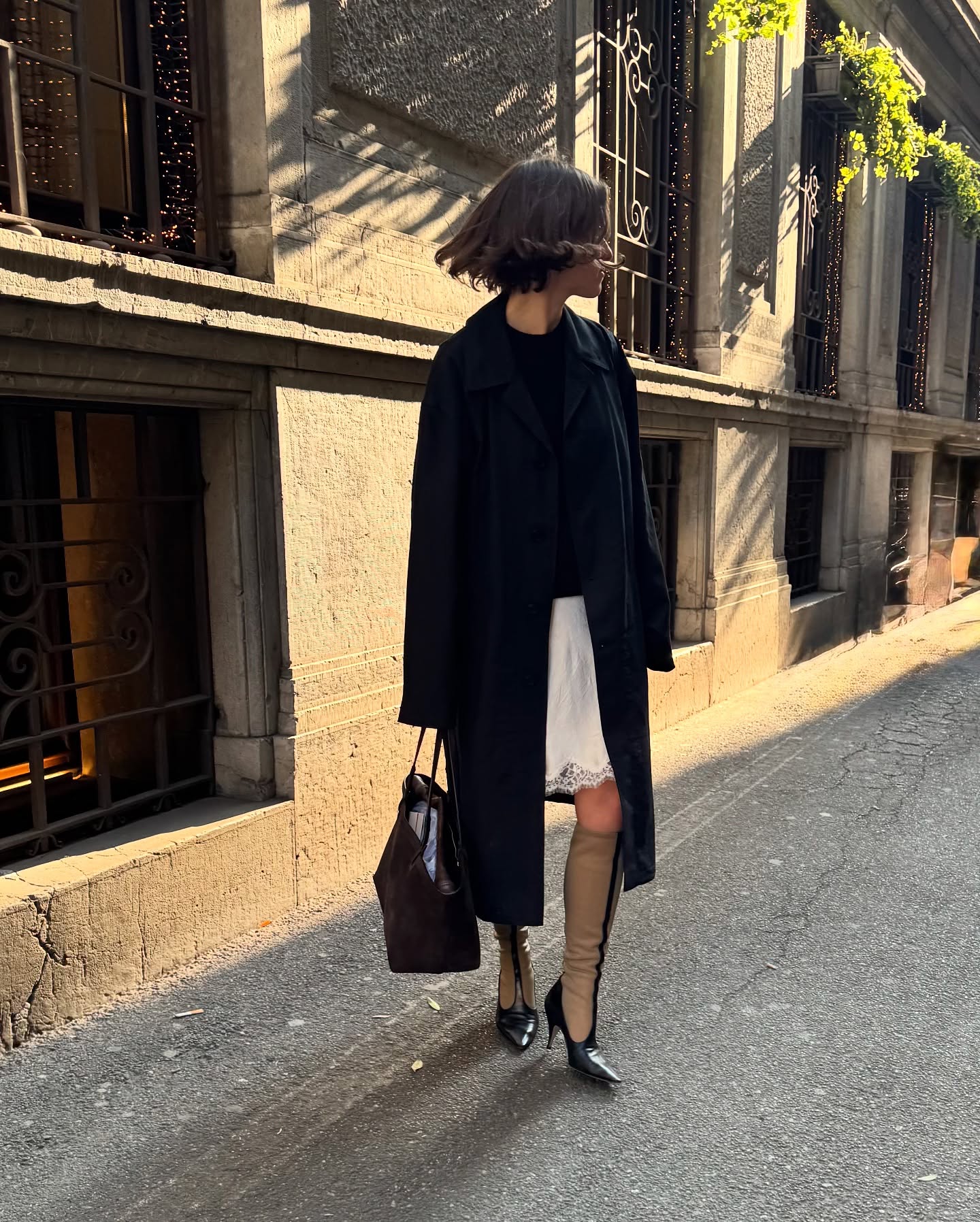 An image of @laurareilly___ wearing a lace-trim slip skirt with a black trench coat and boots from The Row.