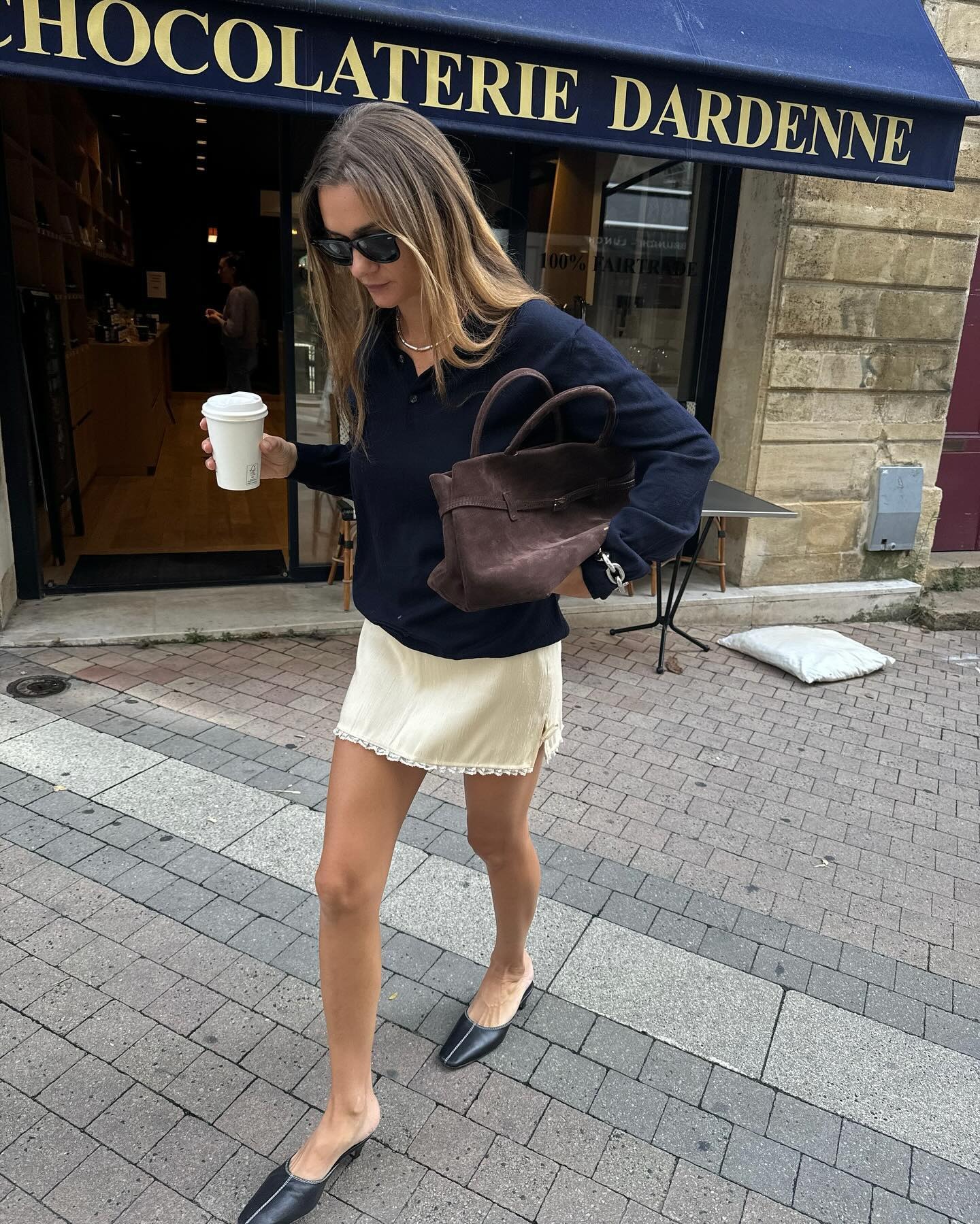 French style influencer Anne-Laure Mais walking on a Paris sidewalk with a coffee in hand wearing oversize sunglasses, a navy polo sweater, a brown suede bag, an ivory satin skirt with lace hem , and black mule kitten heels