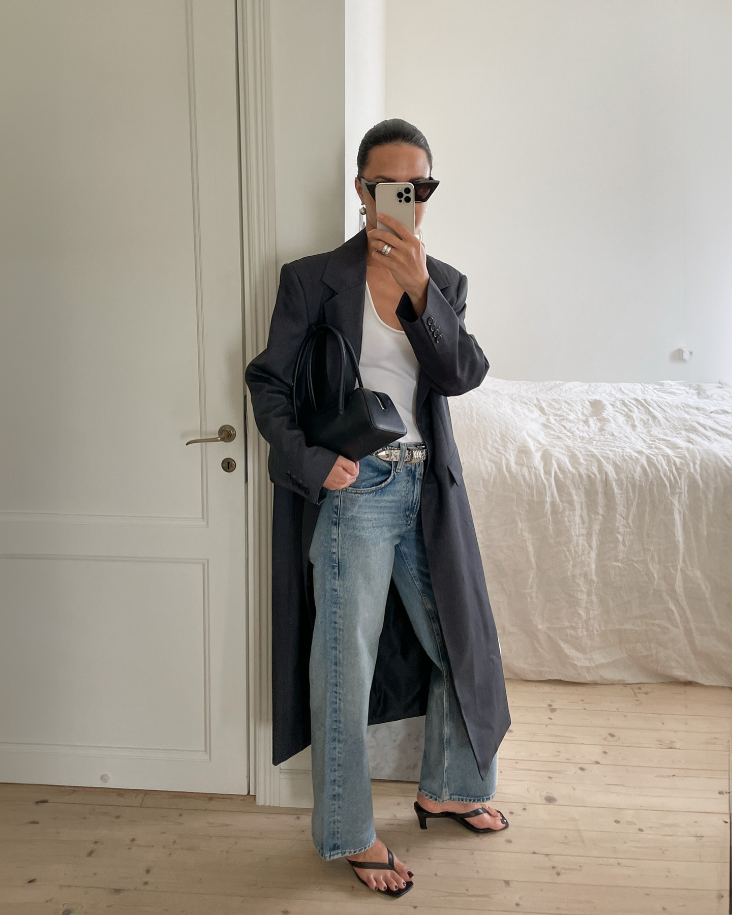 style influencer Johanna Piispa poses for a mirror selfie in her chic home wearing sunglasses, a dark gray maxi coat, white top, black east-west bag, belted jeans, and black flip-flop kitten-heel sandals
