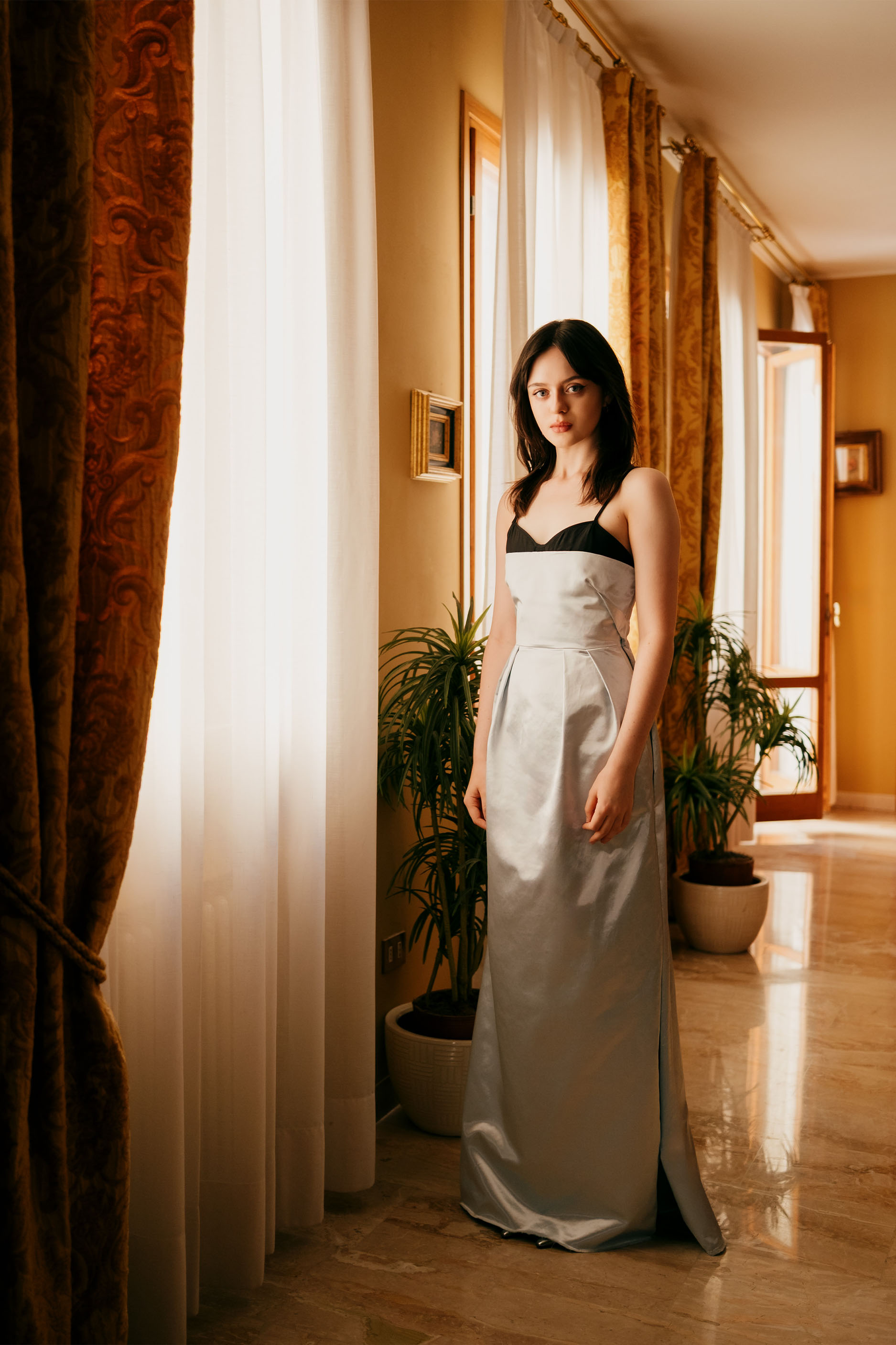 Viola Prettejohn poses for a photo at her Venice hotel. She is standing next to a window wearing a Miu Miu silver satin gown with black bra detailing.