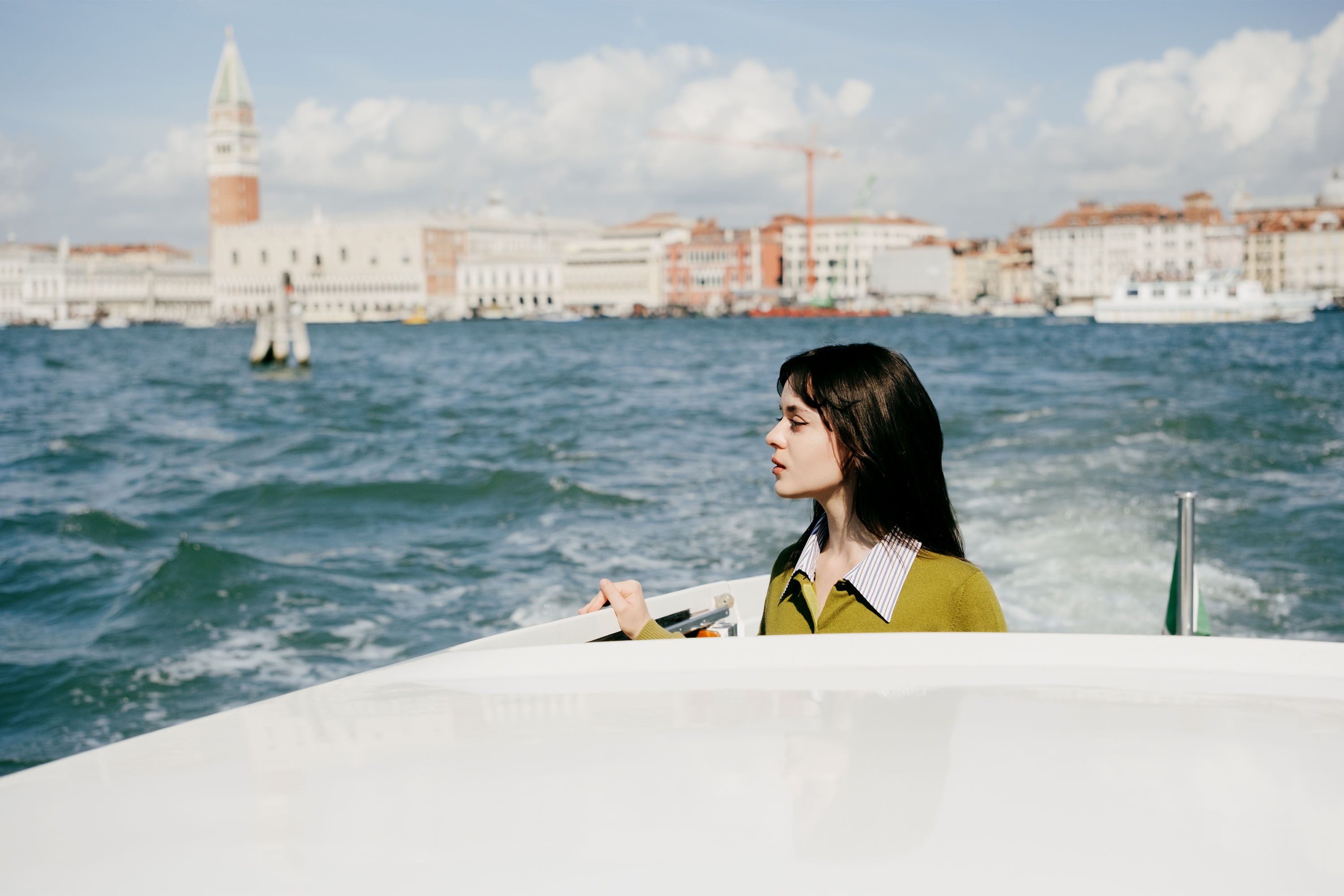 Actress Viola Prettejohn riding in a boat at the Venice Film Festival. She is wearing an olive green cardigan by Miu Miu and the Venice skyline is in the background.