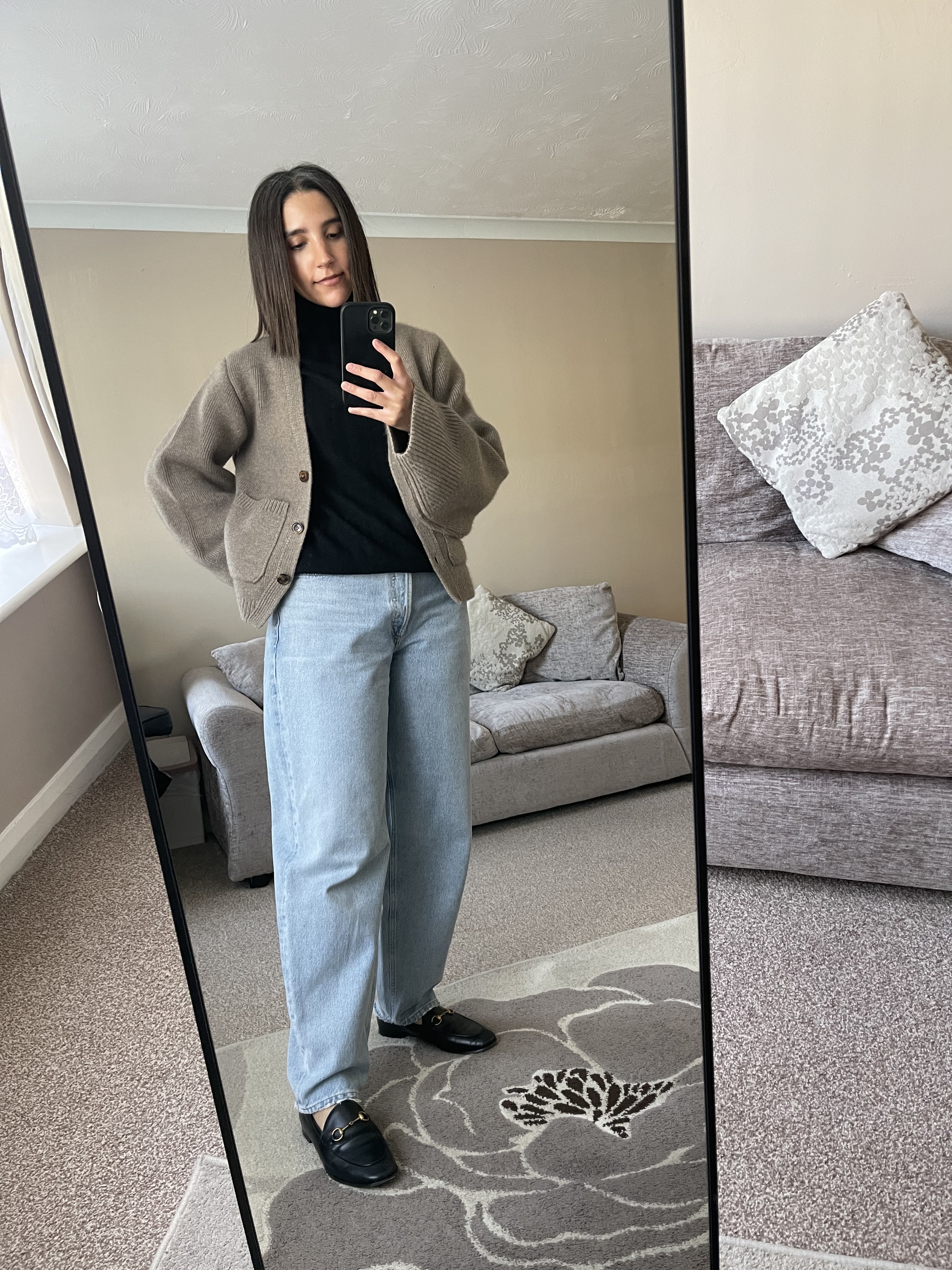 Woman poses in a mirror wearing baggy blue jeans with a black top, grey cardigan and black loafers