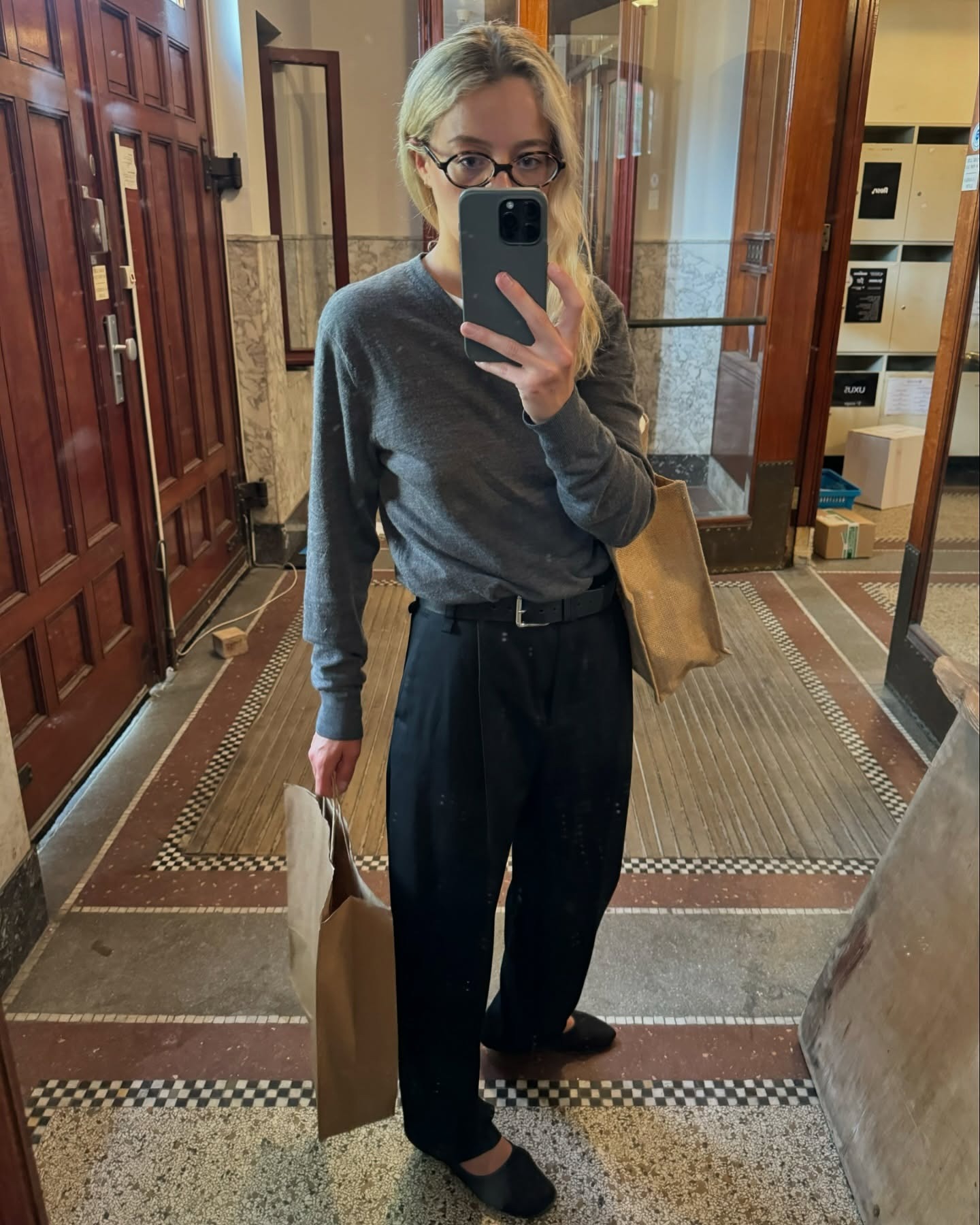 A woman wearing a gray v-neck sweater and a pair of black trousers while carrying shopping bags.