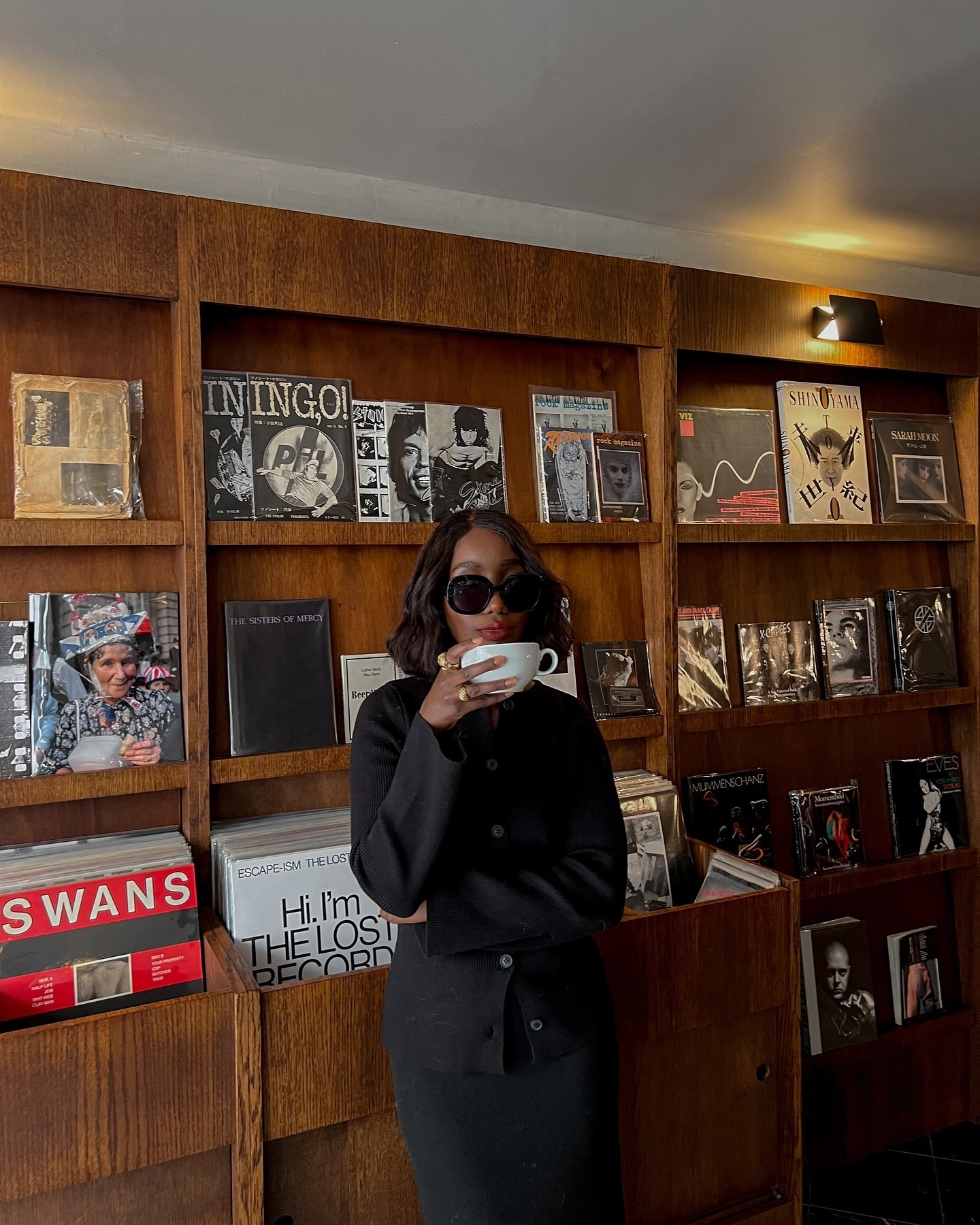 A woman leaning against a record shelf in a black button-down and skirt drinking coffee.