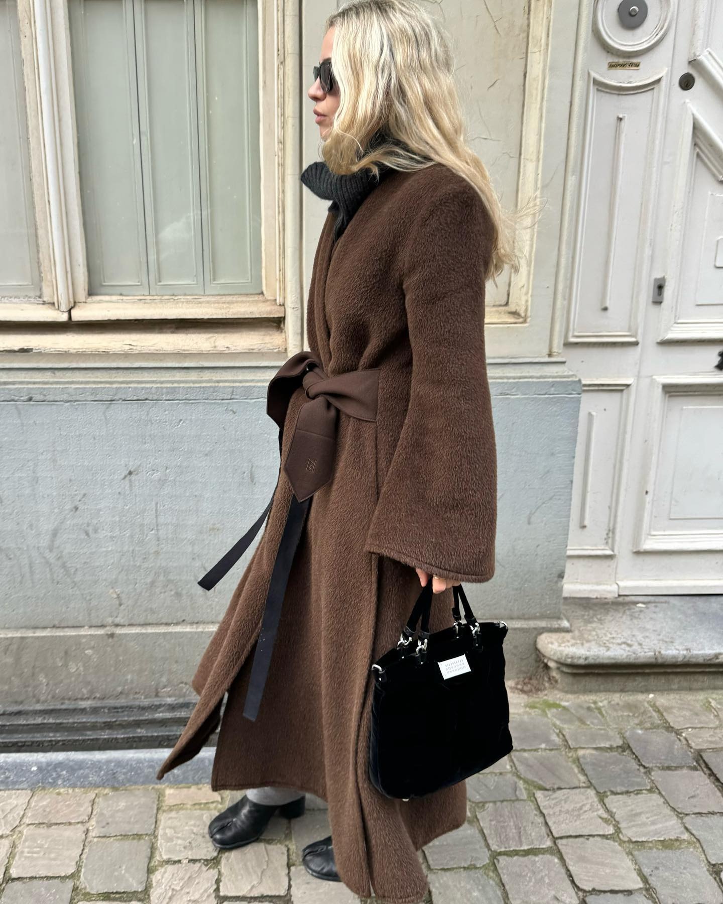 A woman walking down a city street wearing a warm, long coat, a gray turtleneck, black shoes, and a black purse.