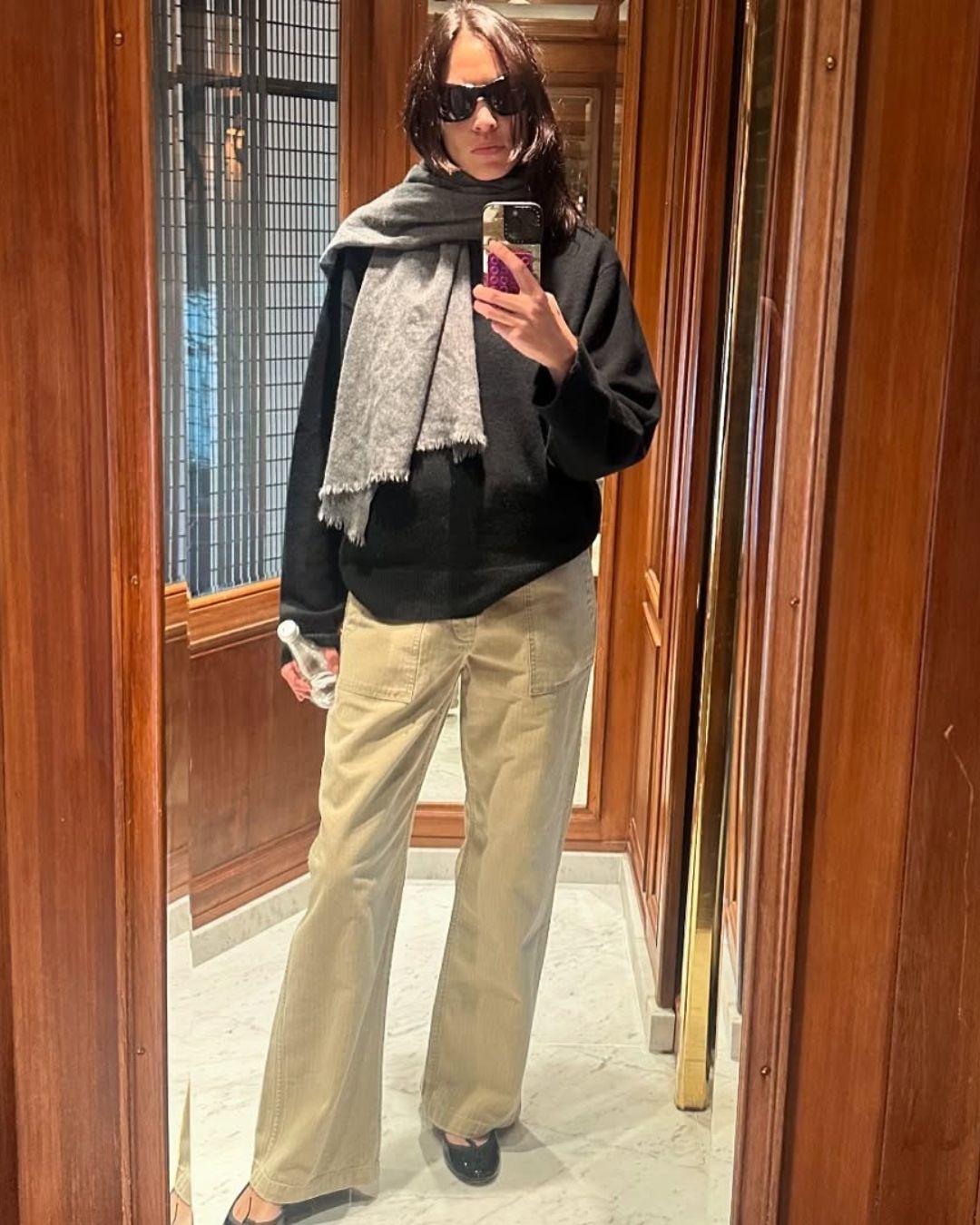 Alexa Chung takes a mirror selfie wearing a black jumper with beige jeans and a grey scarf.