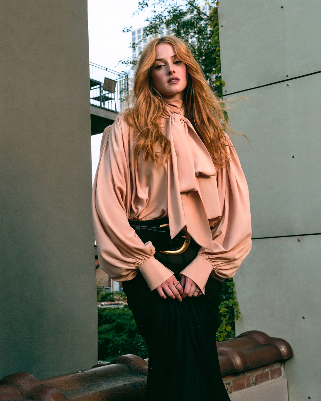 Anna Van Patten wears a blouse and a skirt with a big gold belt.