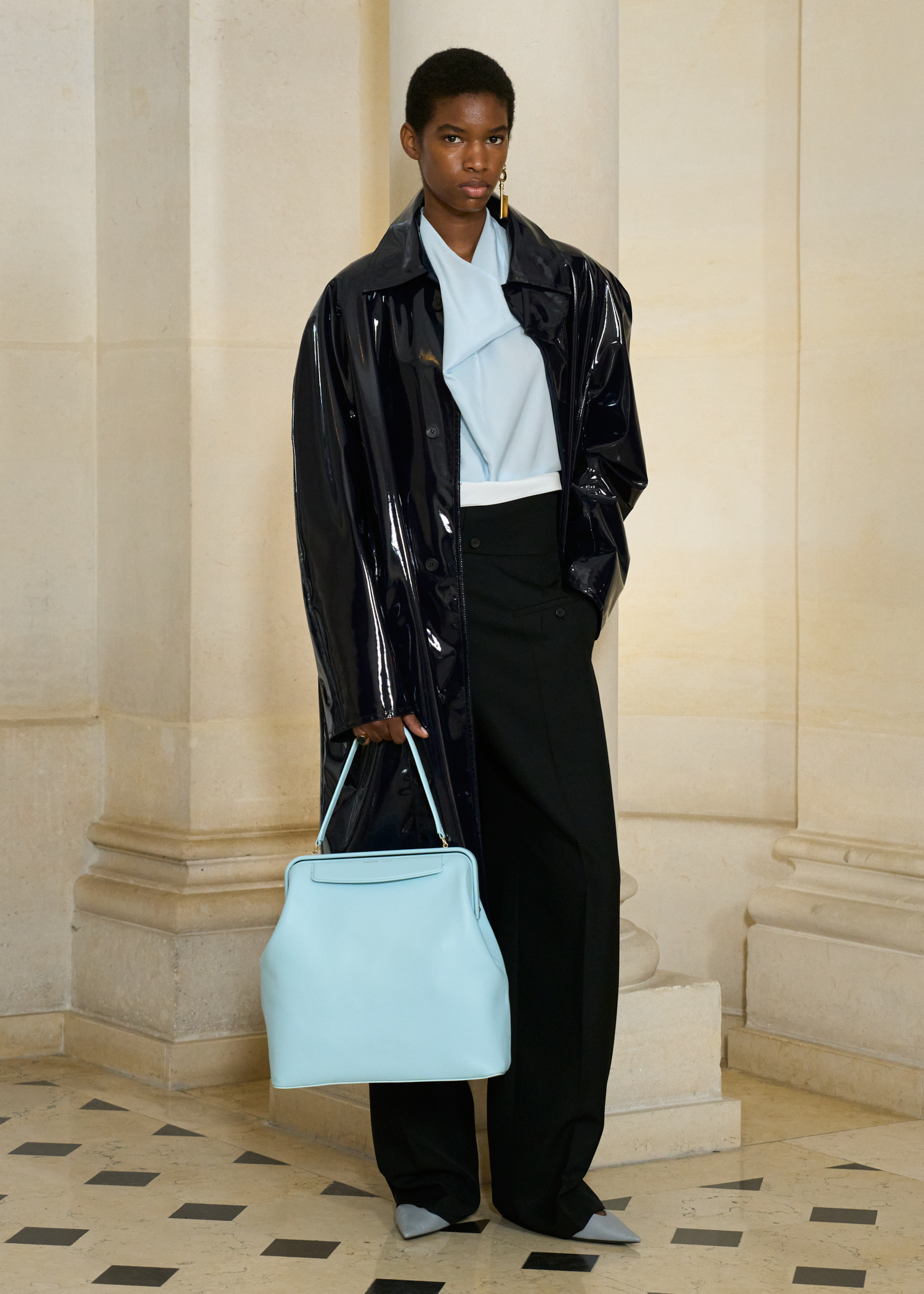 A model in the Carven S/S 26 show wearing a patent-leather coat, a light-blue halter top, trousers, and a light-blue kisslock bag.