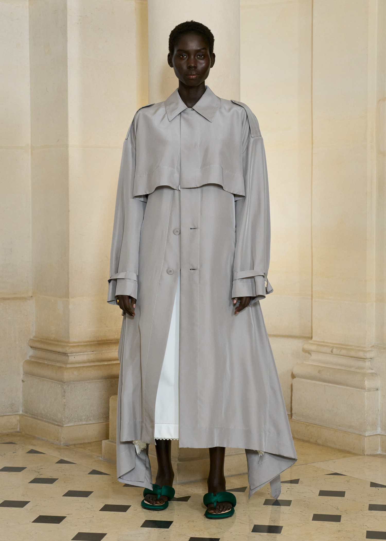 A model in the S/S 26 Carven show wearing a gray trench coat with green padded flip-flops.