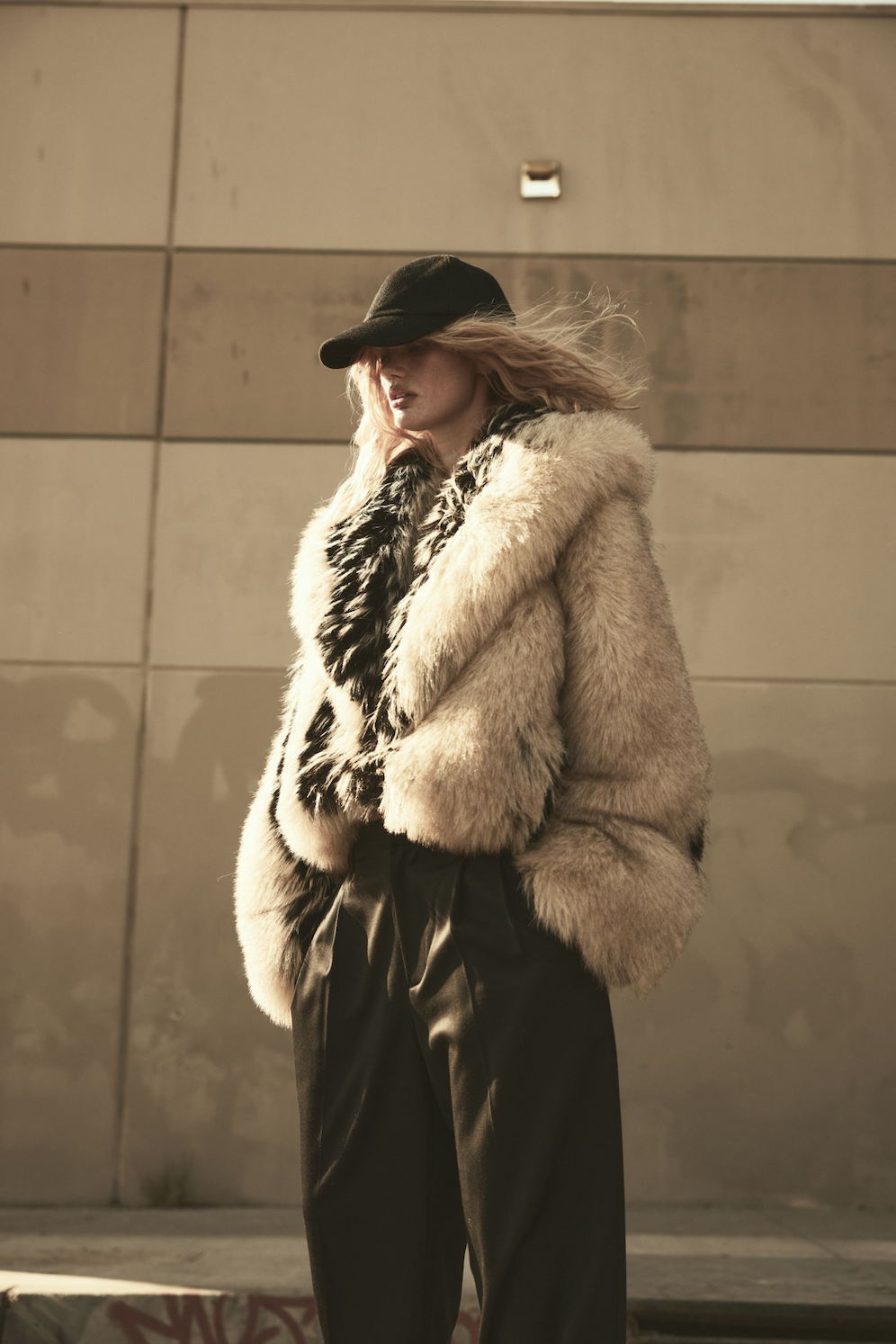 A woman wears a hat, fur coat, and trousers.