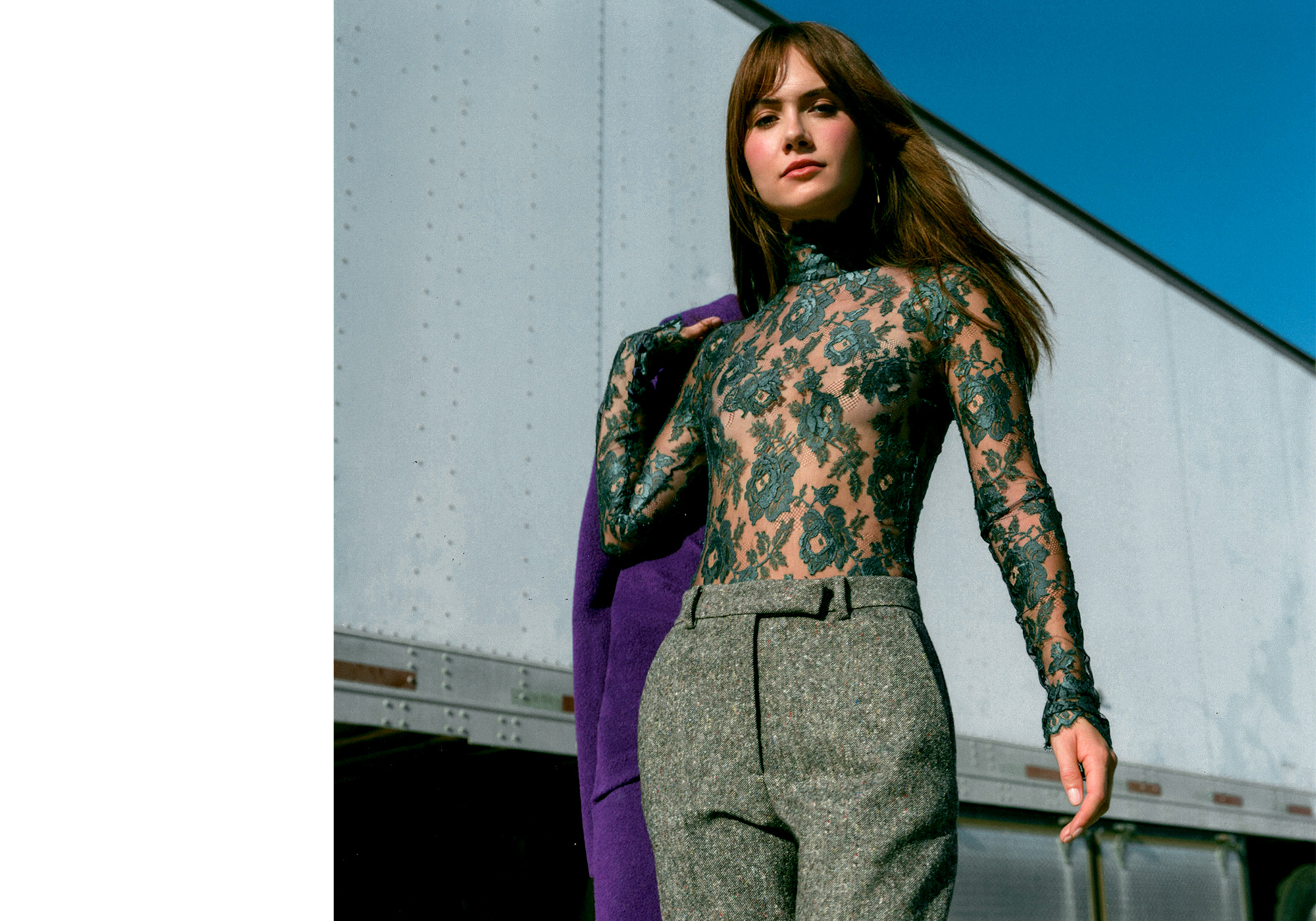 Emilia Jones for WWW&#039;s fall Ones to Watch portfolio. She is standing next to a white semi truck holding a purple coat and wearing a green lace high-neck top and gray trousers.