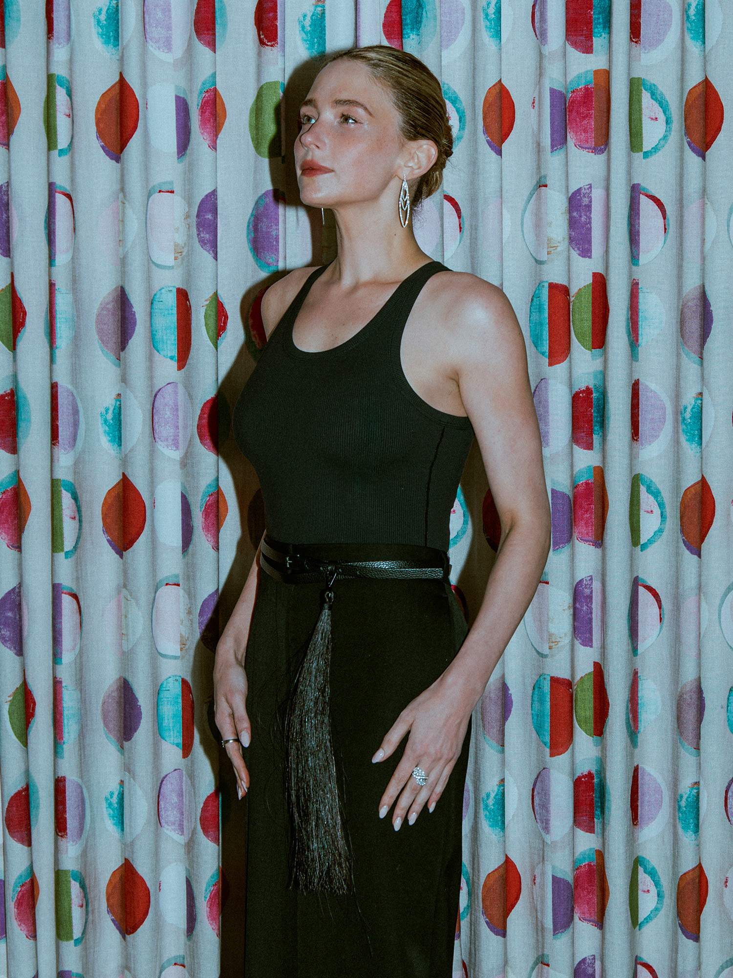 Haley Bennett poses in front of curtains with a colorful circular print. She is wearing a black tank top and maxi skirt with horse hair belt and diamond earrings.