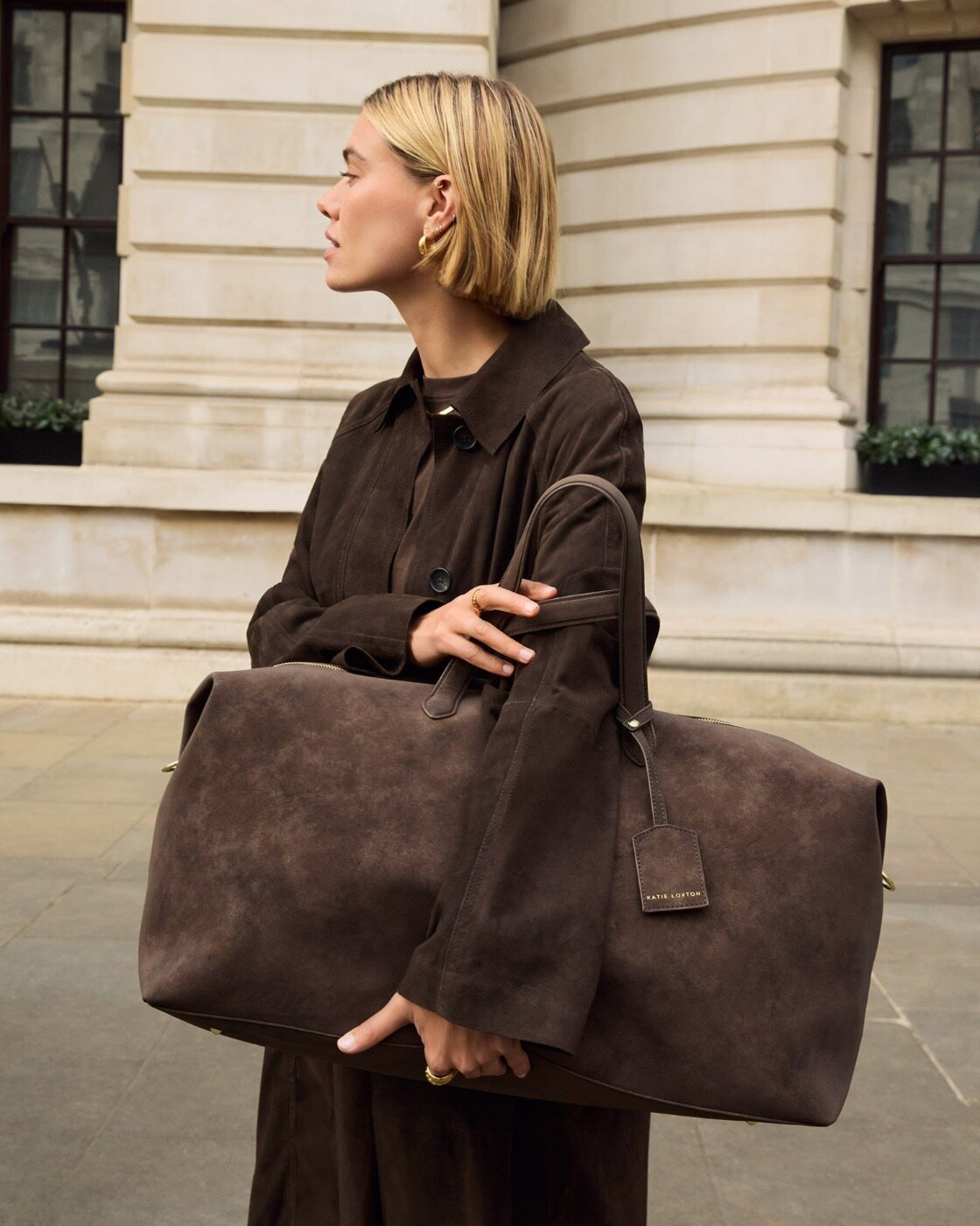 A woman wears a brown suede jacket and brown suede Katie Loxton weekend bag