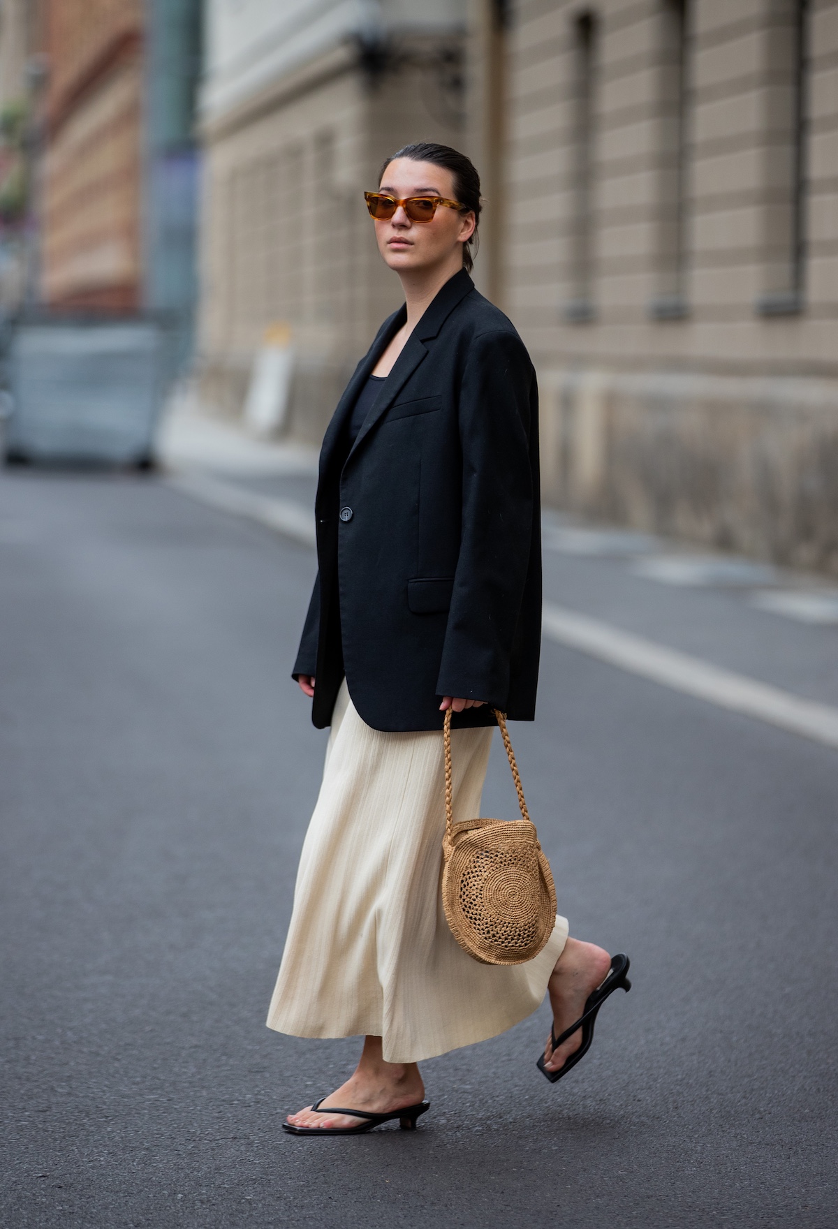 a street style shot of a woman wearing brown sunglasses, an oversize black blazer, a maxi skirt, straw round bag, and black kitten-heel sandals