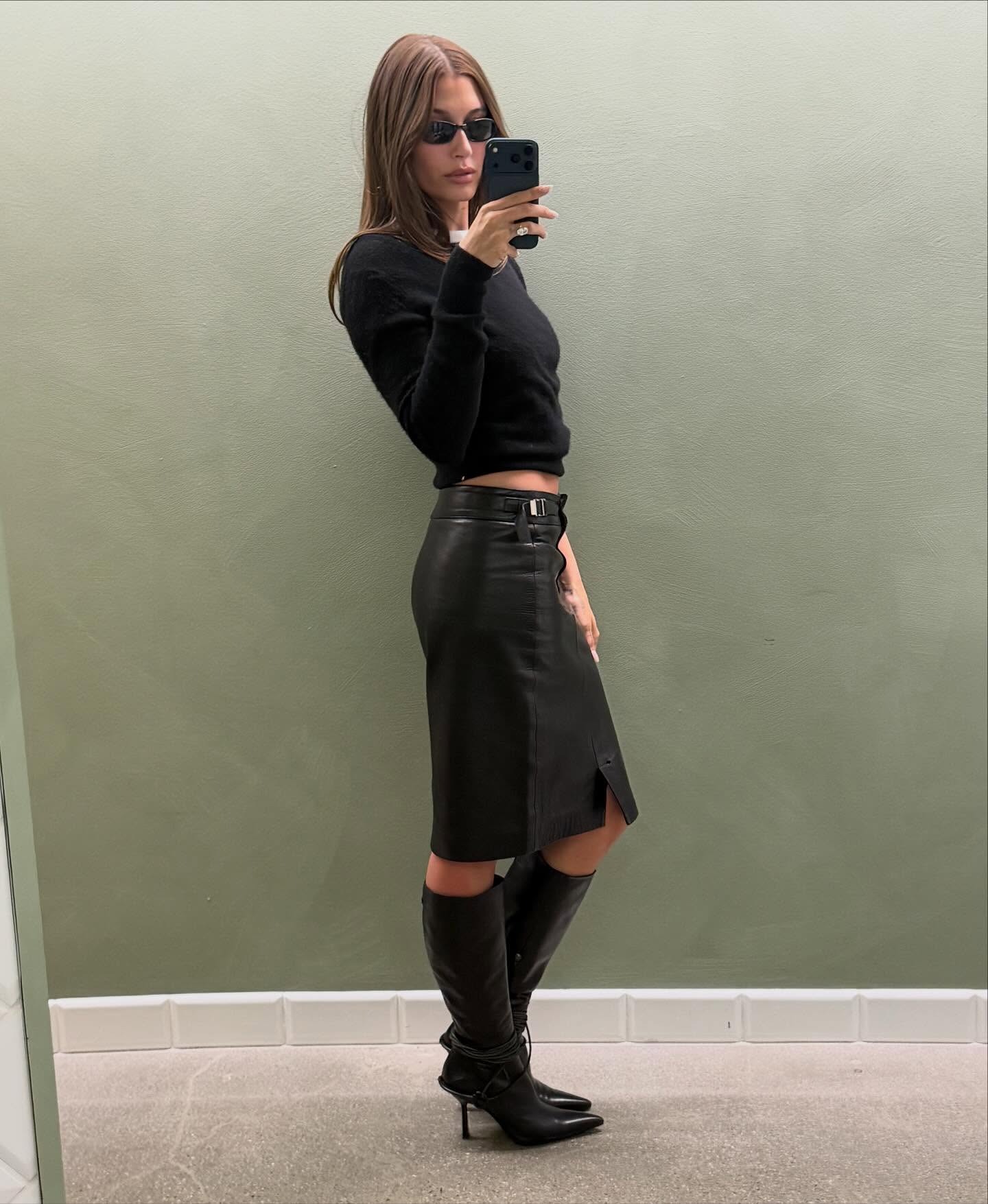 Hailey wears a black sweater, black leather pencil skirt, and black leather knee-high boots.