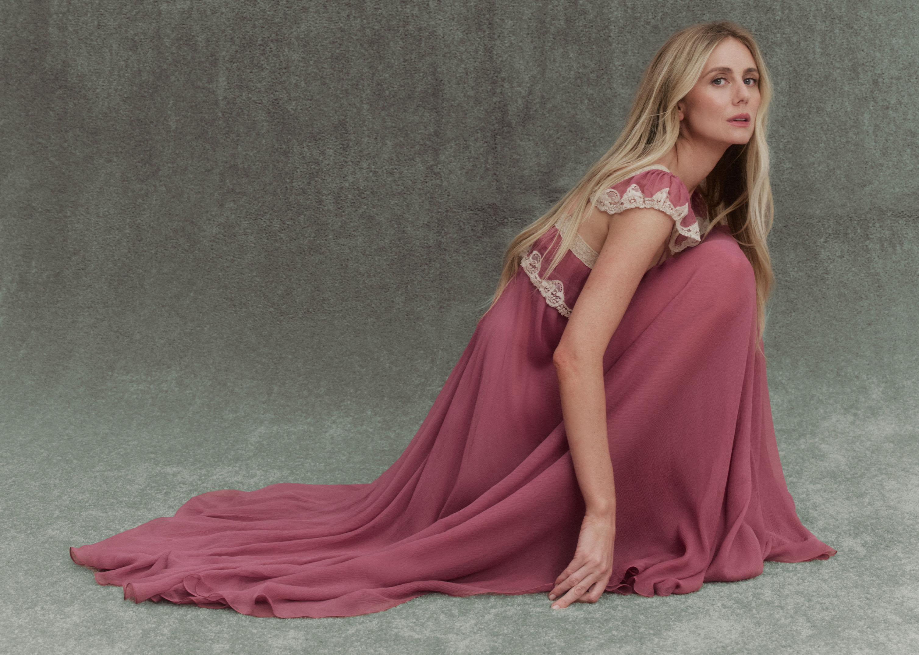 Justine Lupe crouched in front of a gray background wearing a rose-colored Chloe dress with lace.