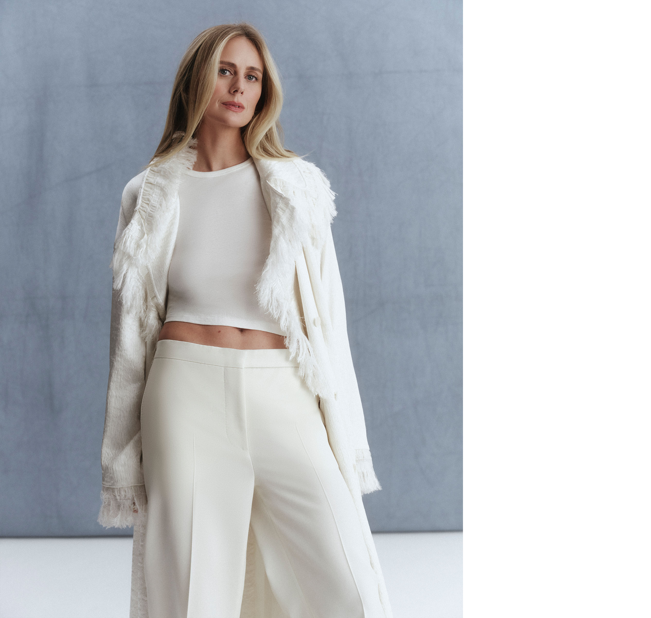 Justine Lupe standing in front of a gray background wearing a white fringed Fforme coat, a t-shirt, and trousers