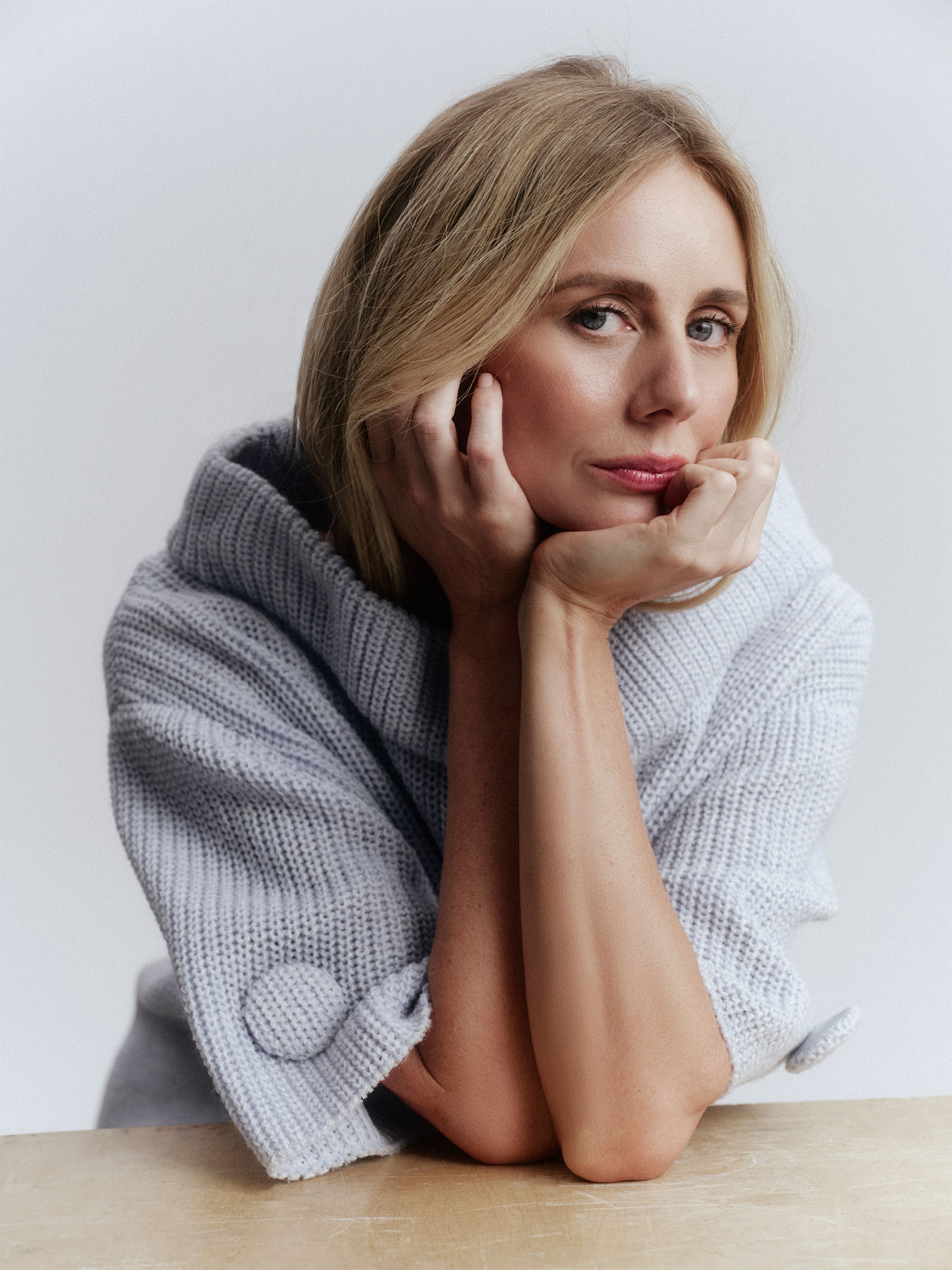 A close up shot of Justine Lupe with her elbows on a table and her chin in her hands wearing a light gray knit Prada dress.