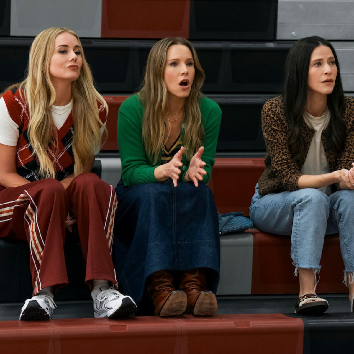 Kristen Bell and co-stars sitting on the bleachers. She&#039;s wearing a a green cardigan, a denim skirt and brown boots.