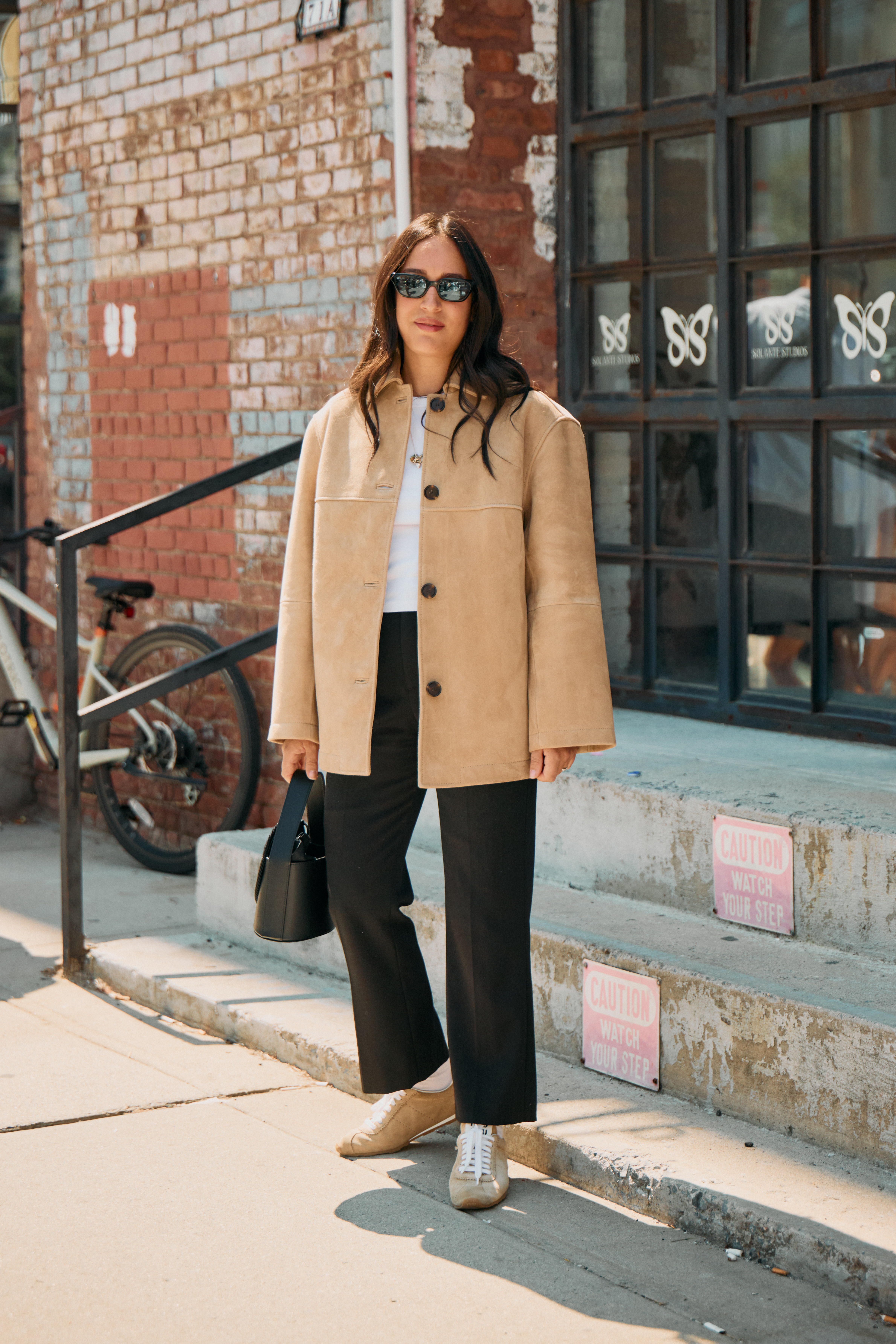 Woman in black trousers, white crewneck tee, low-profile sneakers, and beige suede overcoat.