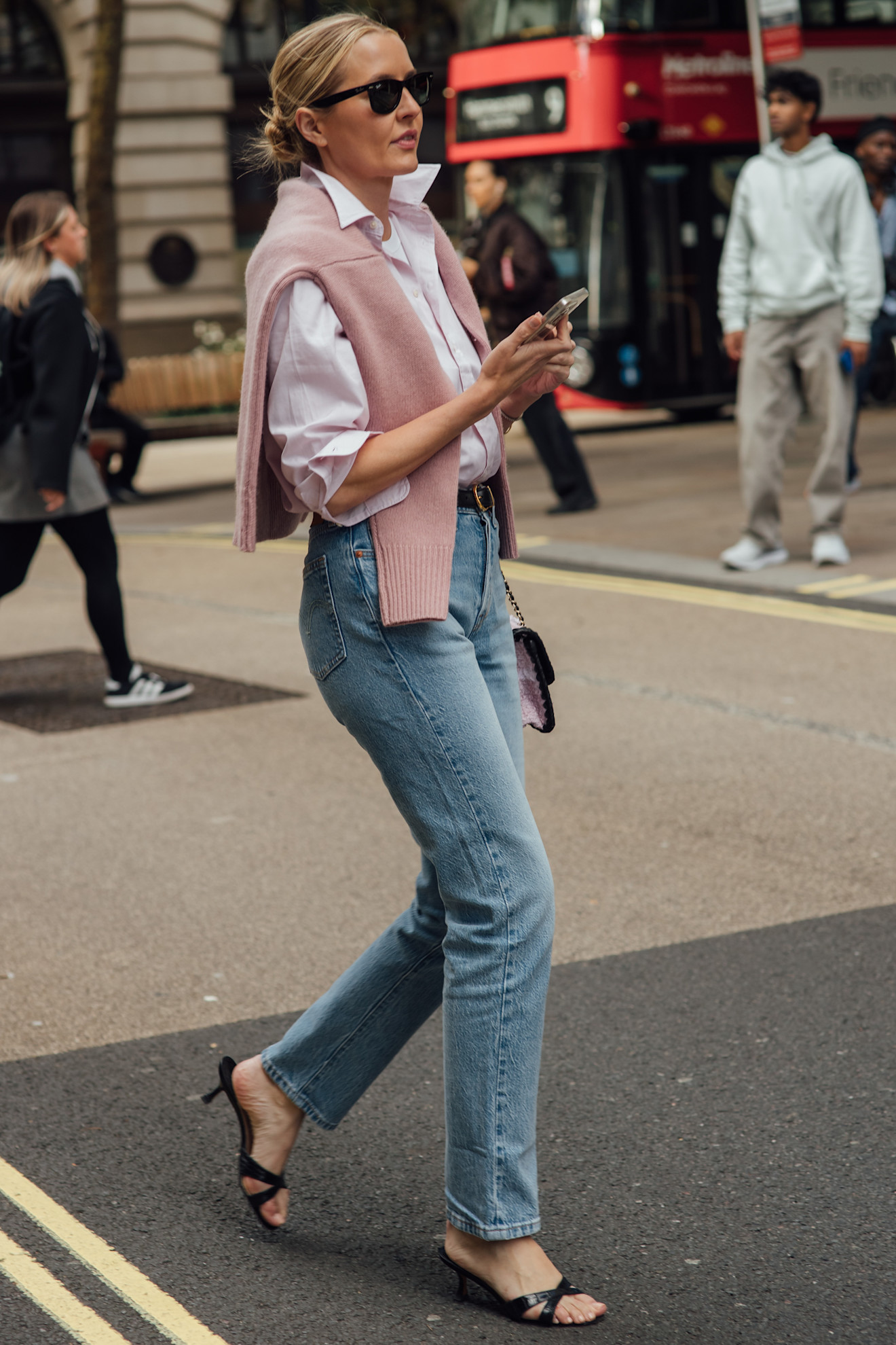 A woman in straight-leg jeans, a button-down, and a pink sweater over her shoulder.