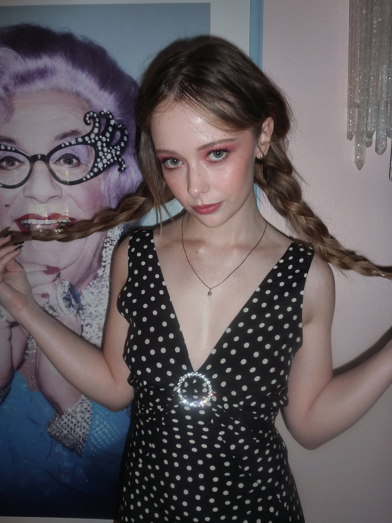 Actress Suzanna Son standing in front of a poster of drag queen. She is wearing a polka dot dress with deep v neck and her hair in braids.