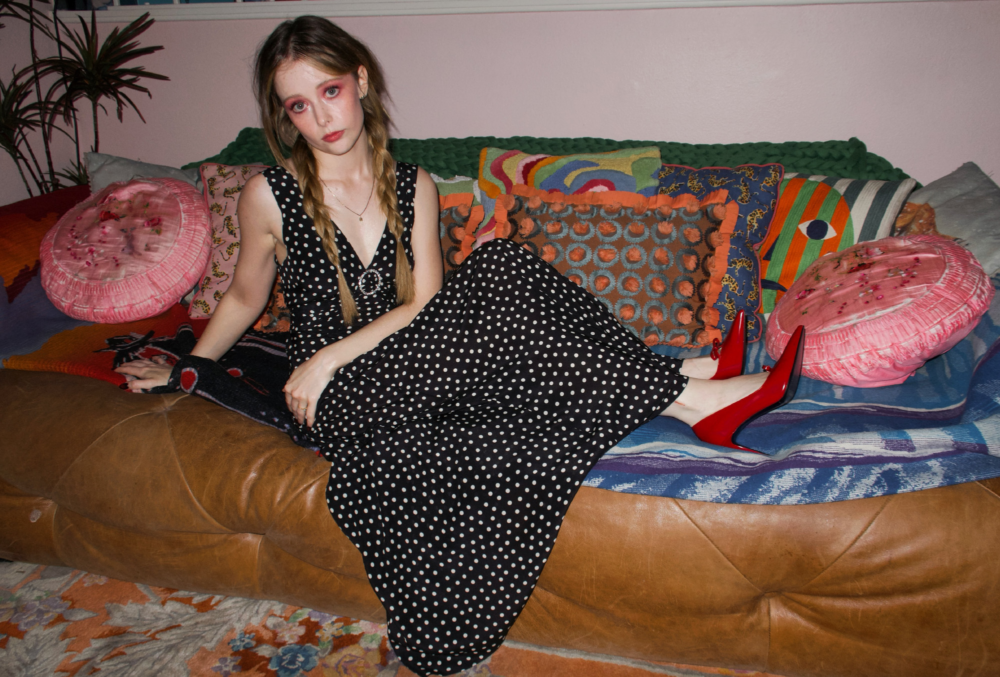 Actress Suzanna Son sitting on a brown leather coach with colorful pillows. She is wearing a polka dot dress with deep v neck, red heels, and her hair in braids.
