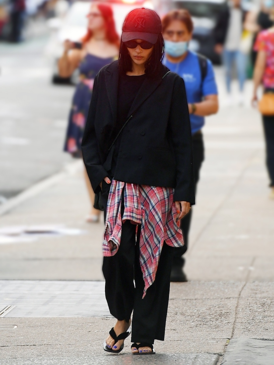 Zoe Kravitz walking in New York City wearing a black Yankees baseball cap, black oval sunglasses, a boxy black blazer, black crossbody bag, a red plaid shirt tied around her waist, ankle-length black pants, and black The Row flip-flop sandals