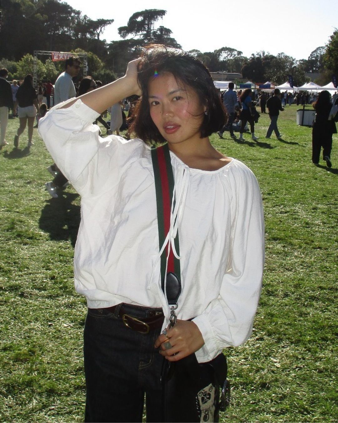 Influencer @alllisonho stands on a grassy field wearing a white balloon blouse with a cross-body bag and dark wash jeans.