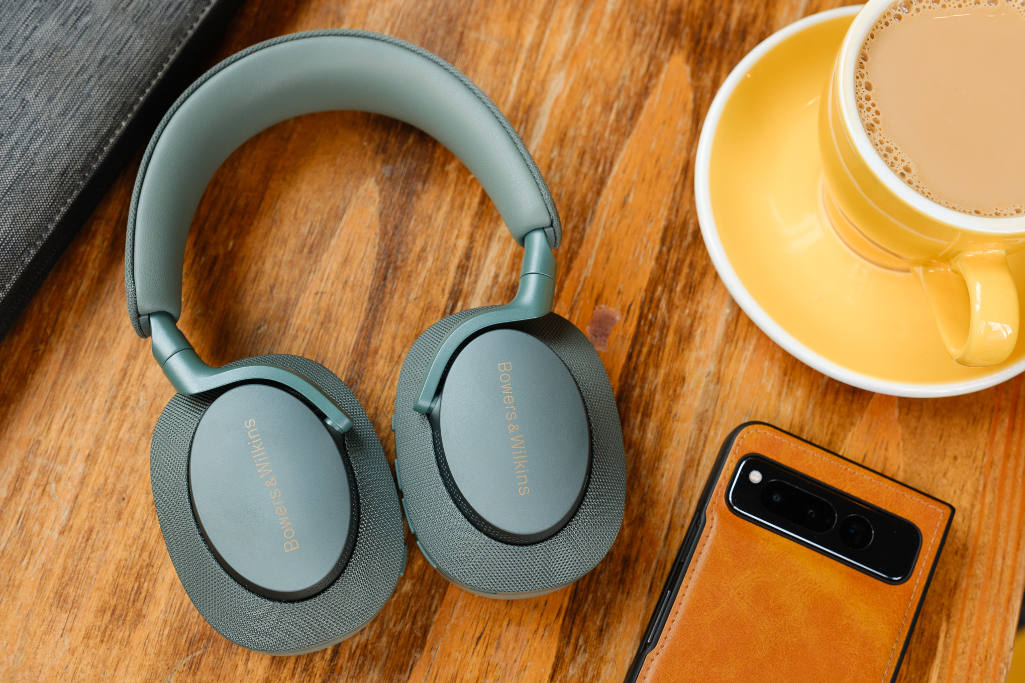 A pair of green Bowers &amp; Wilkins PX7 S2e headphones on a wood table beside a phone and coffee.