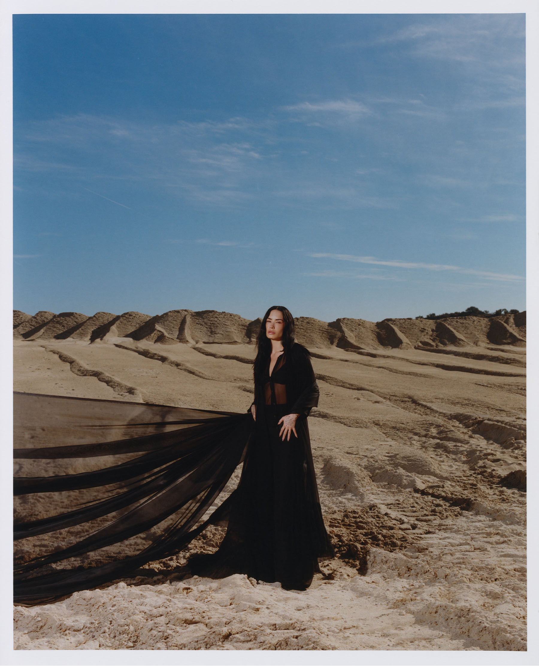 Demi Lovato for Who What Wear&#039;s November cover. Lovato is standing in the middle of a sand dune wearing a black maxi dress. The fabric is flowing off to the side.