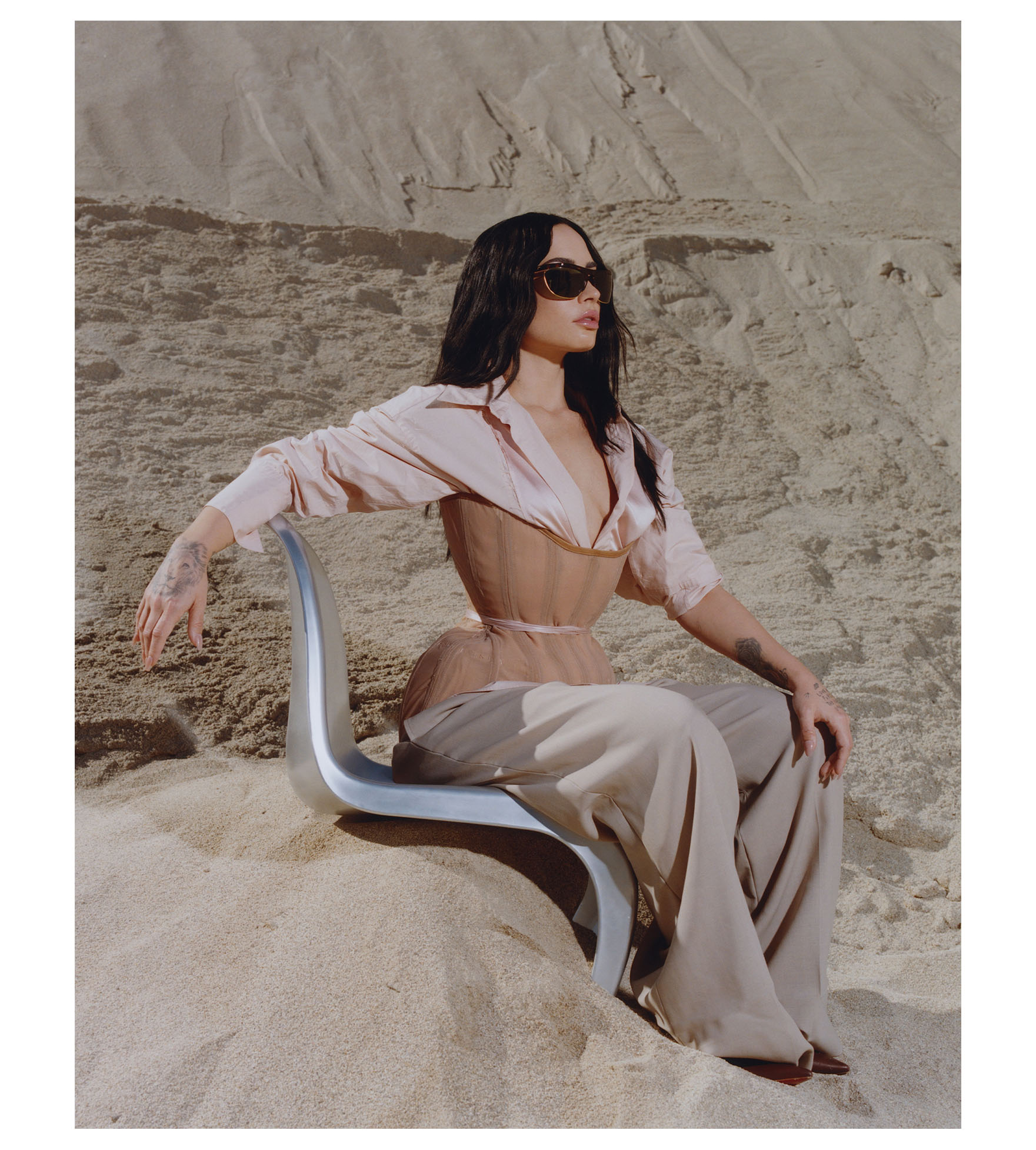 Demi Lovato for Who What Wear&#039;s November cover. She is sitting on a silver chair with her left hand on her knee on a sand dune. She is wearing a pink button-down shirt with a nude corset on top and tan trousers. She is also wearing sunglasses.