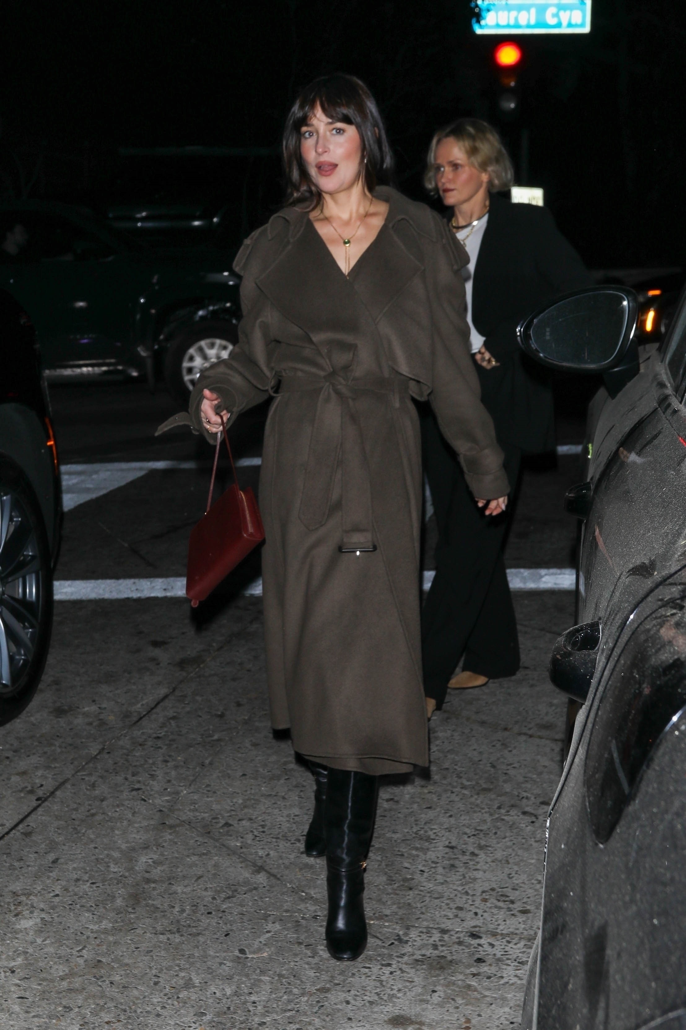 Dakota Johnson walks down the street wearing a dark brown belted coat with black knee-high boots.
