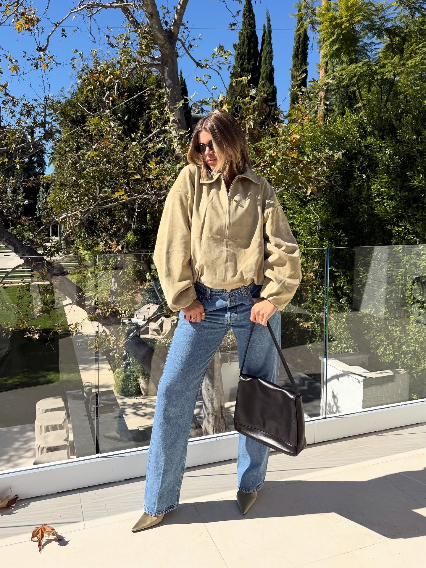 Sofia Richie Grainge poses on a balcony wearing a light beige suede jacket, blue vintage Levi jeans and satin pointe-toe heels. She holds a black leather bag in her hand and wear black sunglasses.