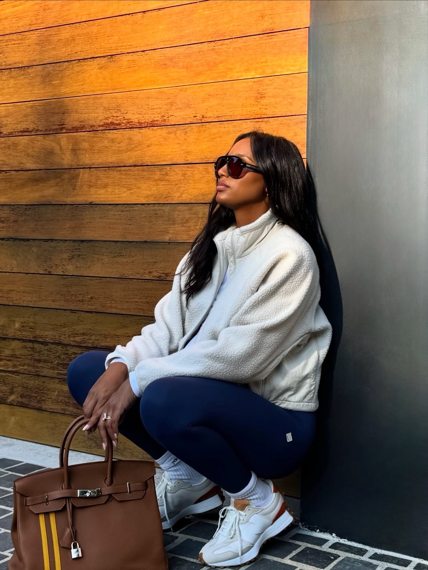 Influencer @jastookes crouches down wearing navy leggings with white New Balance trainers, a white fleece and a brown Hermes bag.