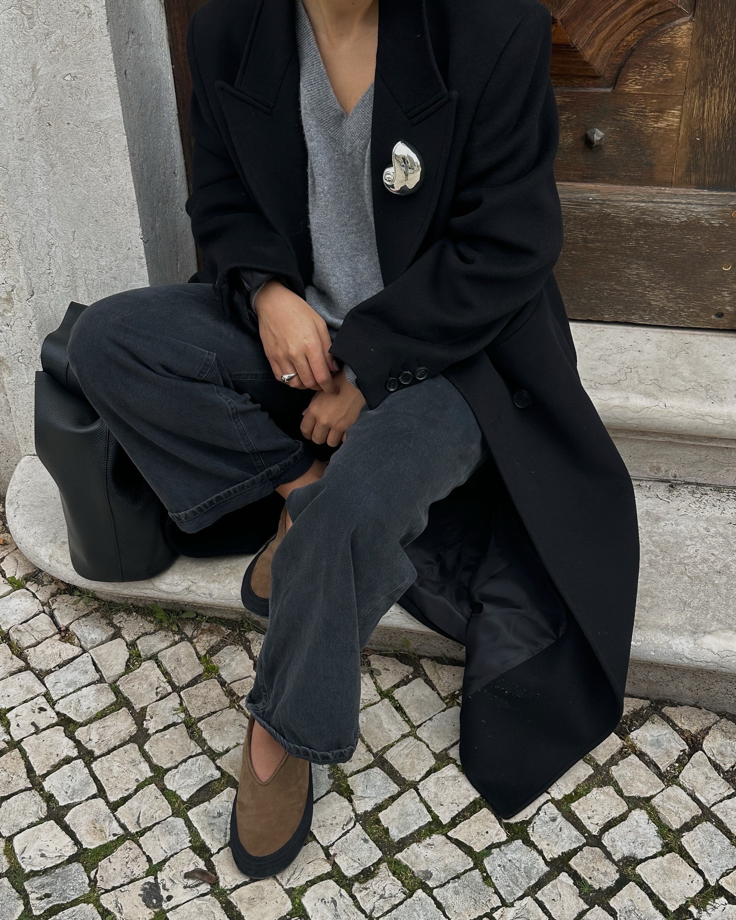Influencer sits on steps outside wearing black jeans with suede flats, a black coat and a silver brooch.