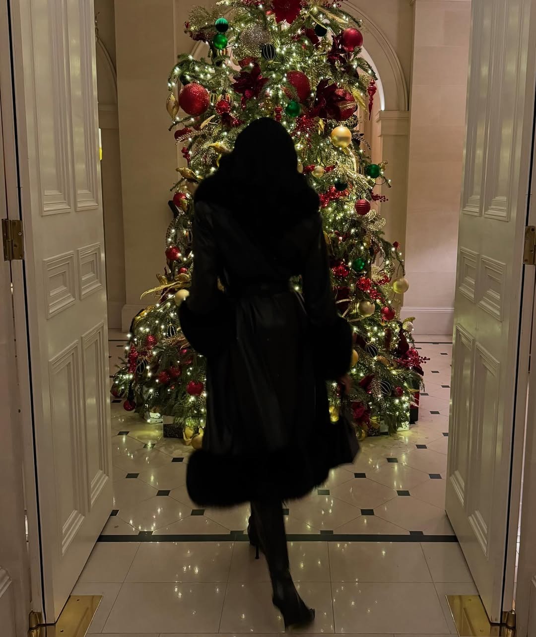 A woman wearing all-black walking in front of a Christmas tree.
