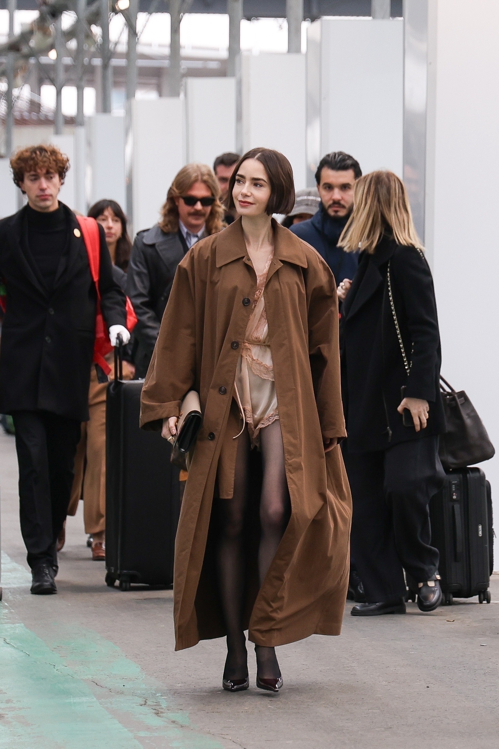 Lily Collins arrives at Gare d&amp;rsquo;Austerlitz in Paris wearing a lingerie romper from YSL with a trench coat, tights, and heels, for Emily in Paris.