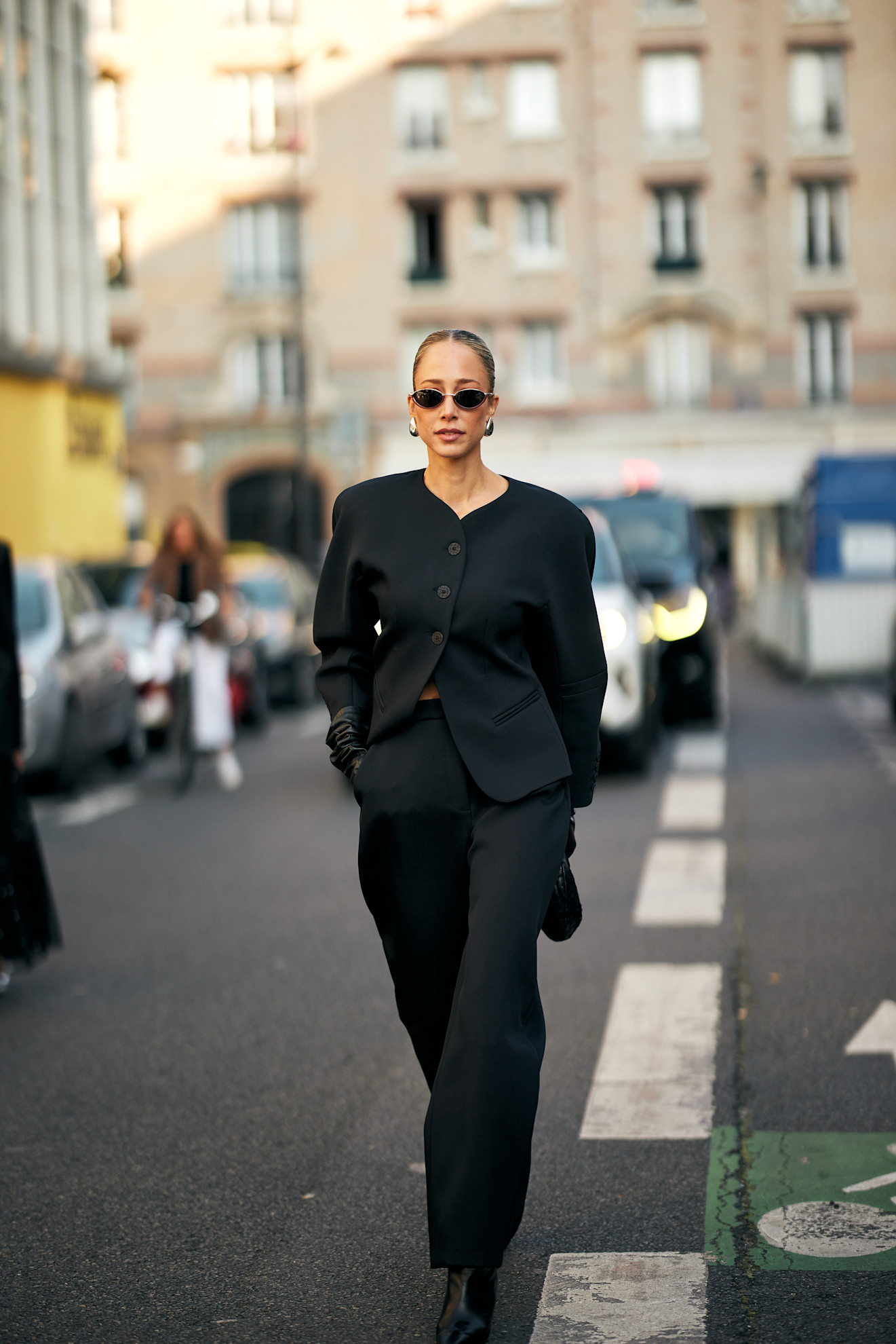 A person wearing black trousers, a collarless blazer, heeled boots, and sunglasses.