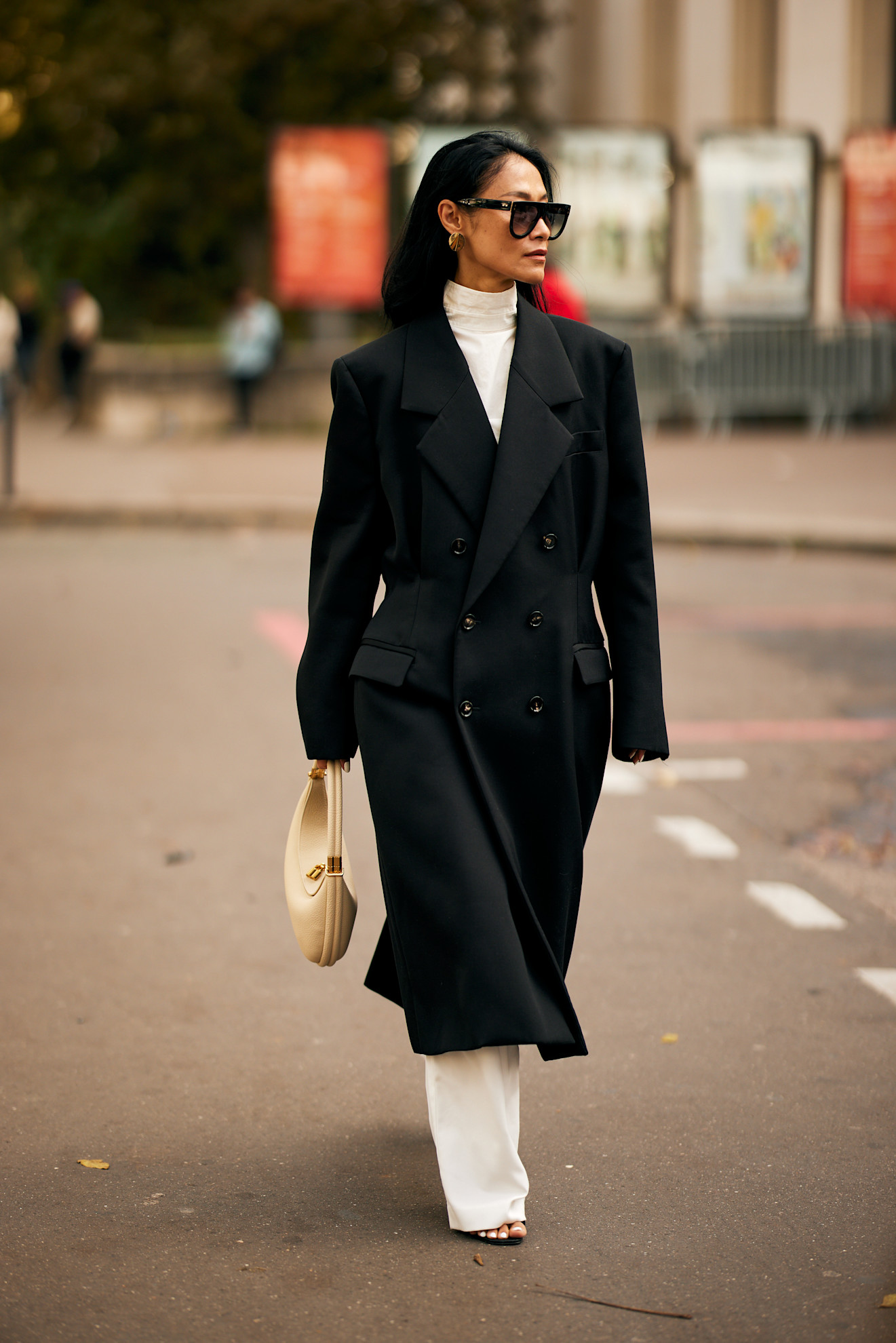 Person wearing wide-leg white trousers, a white turtleneck, an oversized double-breasted coat, a half-moon bag, and sunglasses.