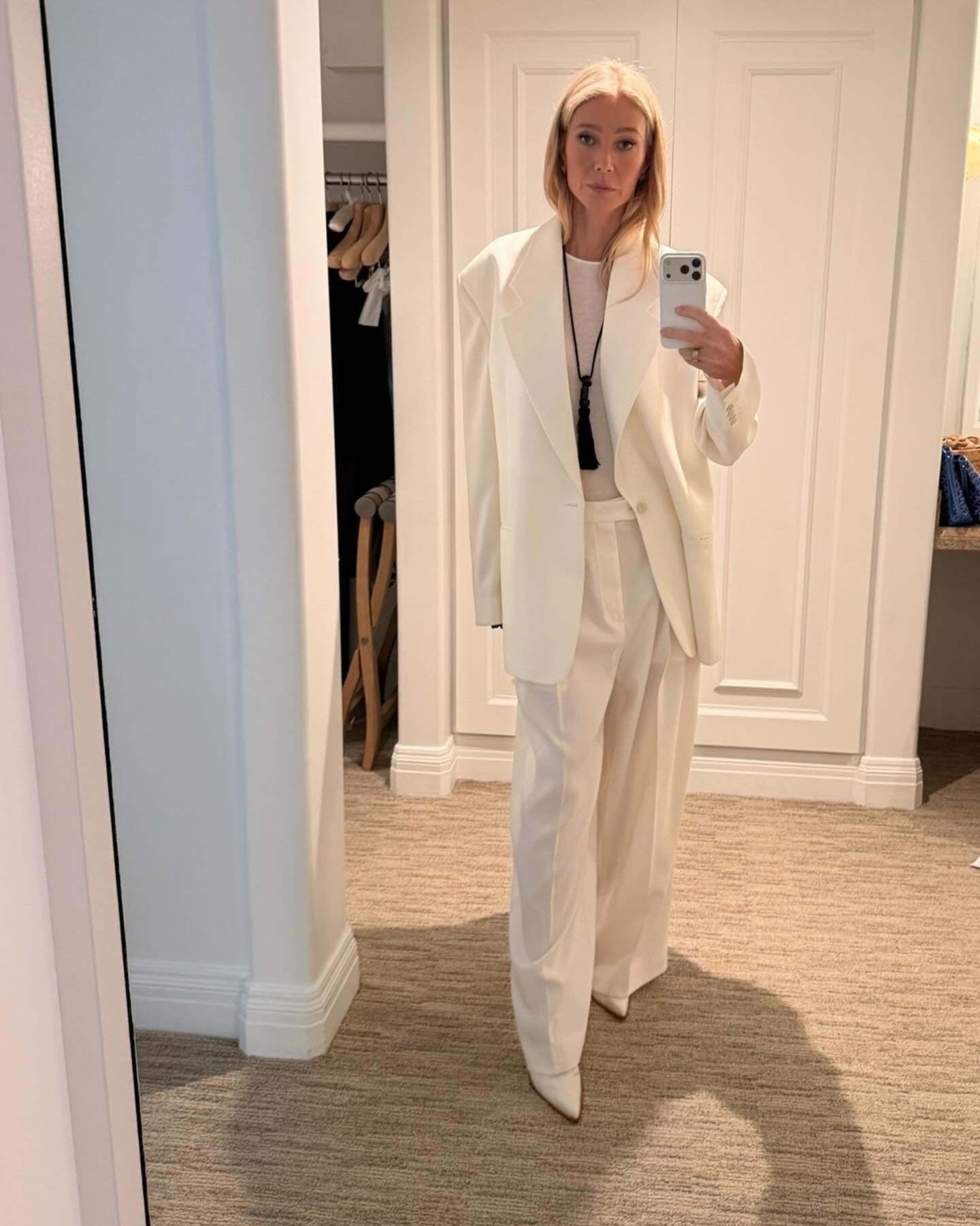 Gwyneth Paltrow takes a mirror selfie wearing a white oversized suit with white pointed-toe heels.