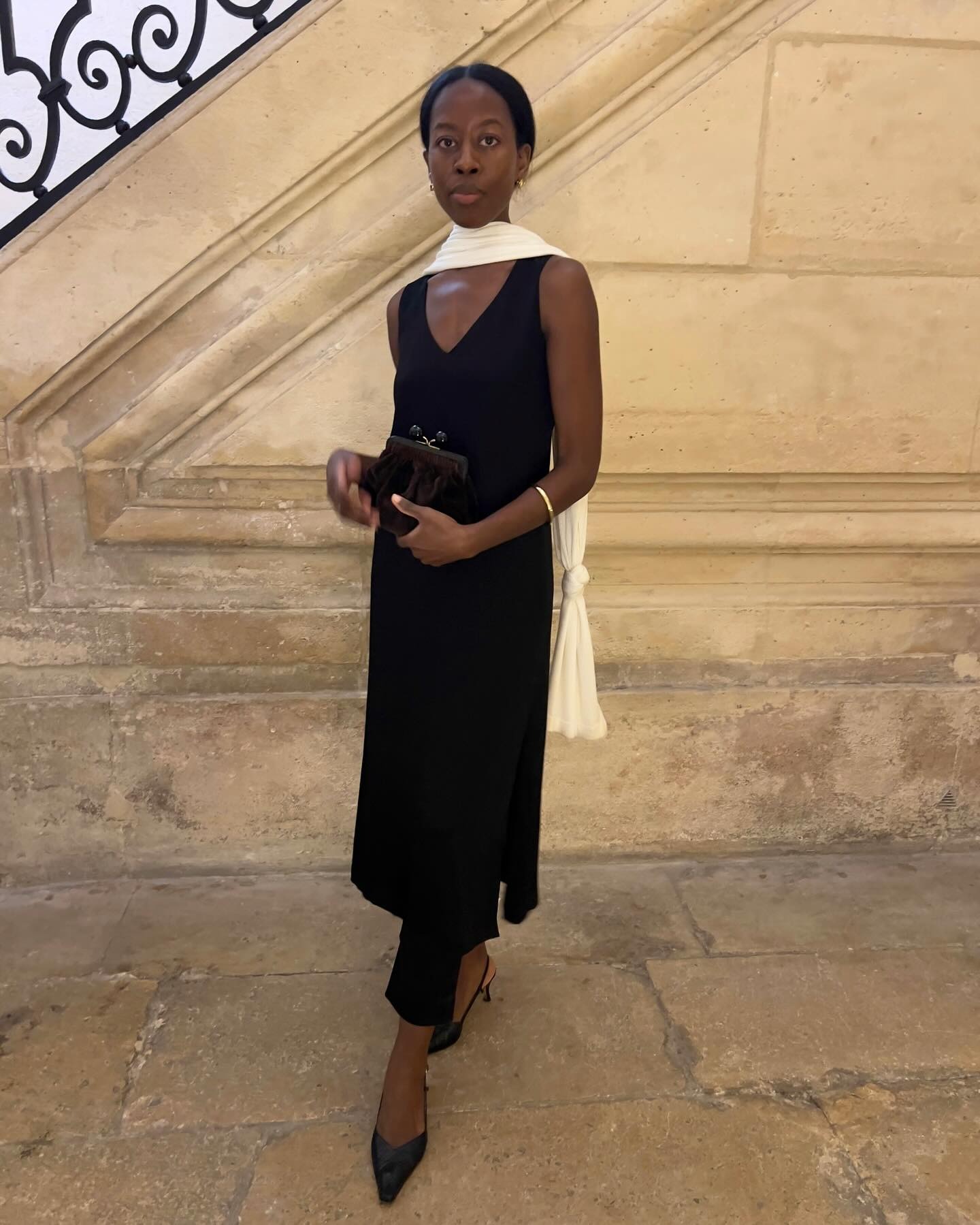 Influencer @sylviemus_ wears a black v-neck dress with black pointed-toe slingbacks and a white scarf.