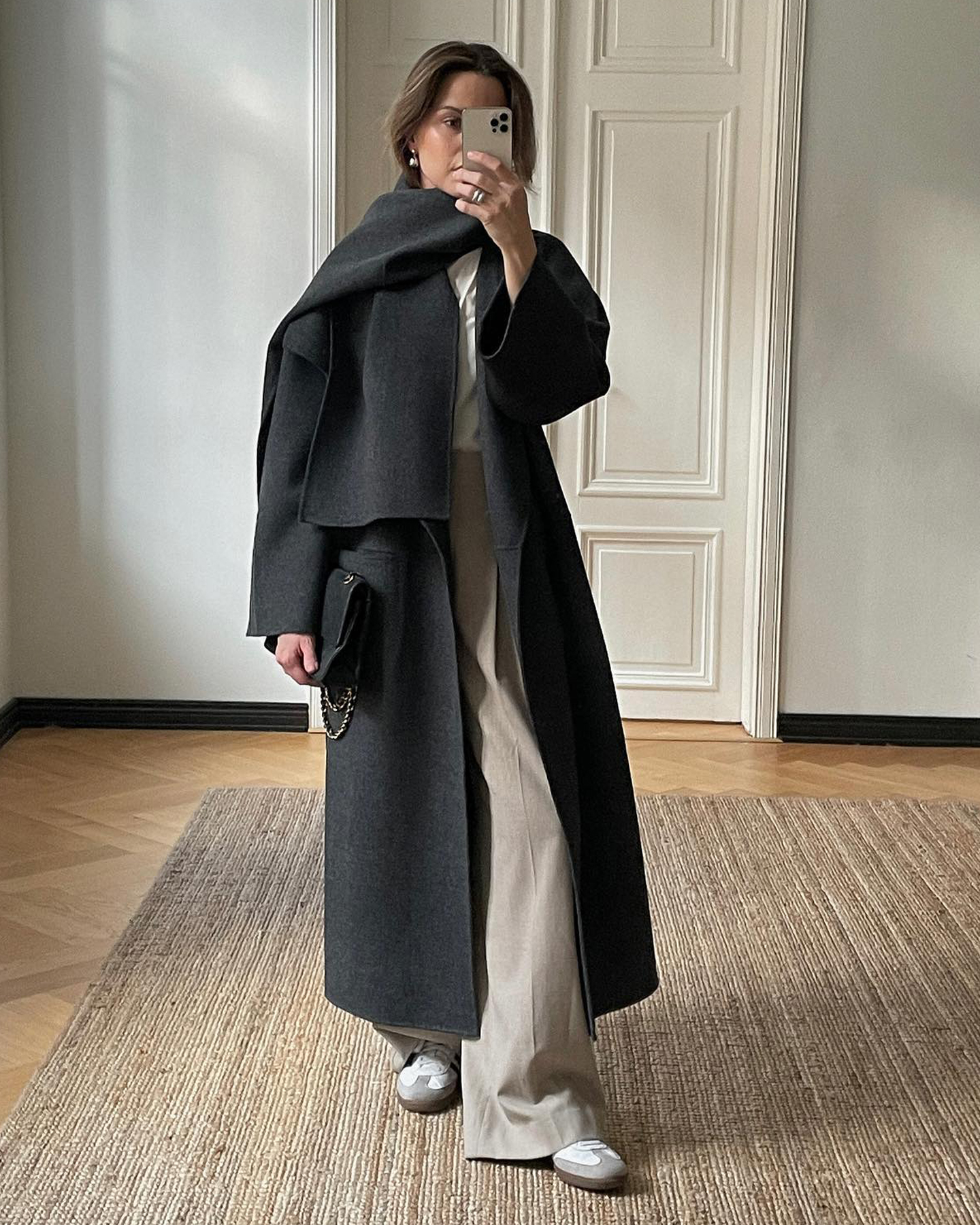 Helsinki style influencer Johanna Piispa poses for a mirror selfie wearing a long charcoal gray scarf coat, a white tee, beige relaxed pants, and Adidas Samba sneakers