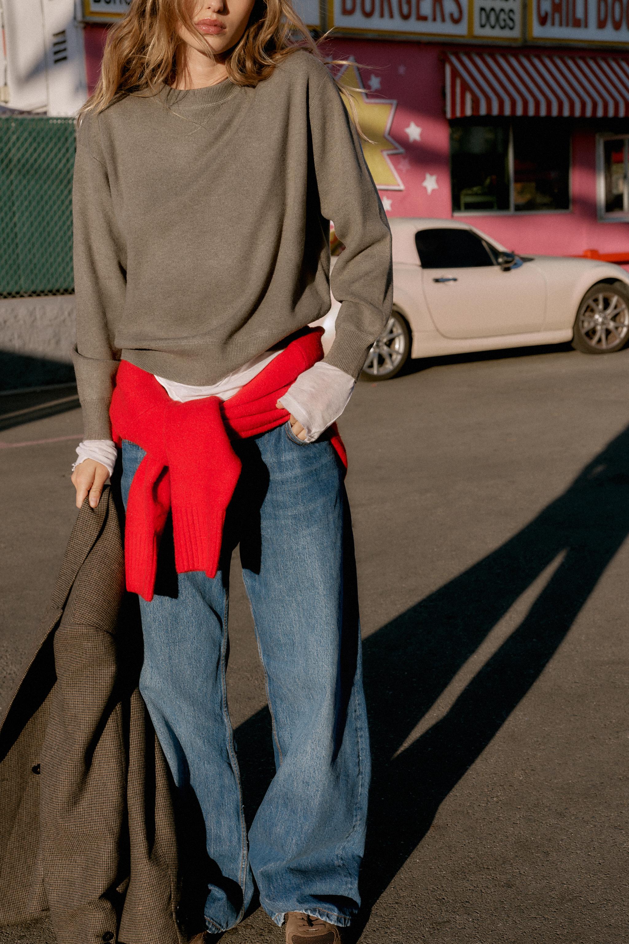 Zara model shot in a chic outfit idea for 2026 with taupe sweater layered over a sheer white long-sleeve top, a red sweater around the waist, relaxed jeans, and brown suede sneakers