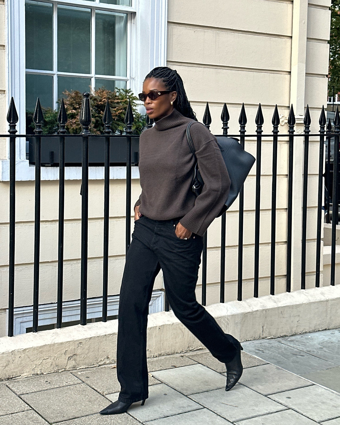 British style influencer Marilyn Nwawulor-Kazemaks walks on a London sidewalk wearing a chic fall and winter outfit with black rectangular sunglasses, a brown turtleneck sweater, The Row bag, black jeans, and the kitten-heel boots trend for 2025