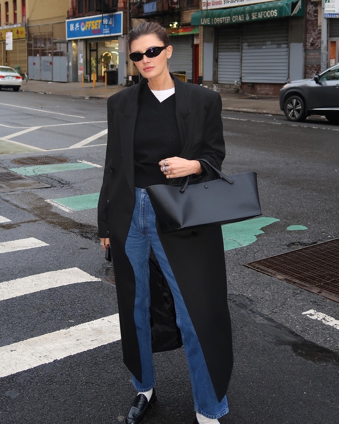 NYC based style influencer Coco Schiffer poses on a New York City crosswalk wearing black sunglasses, a black maxi coat, black V-neck sweater layered over a white tee, The Row bag, straight-leg jeans, white socks, and black loafers