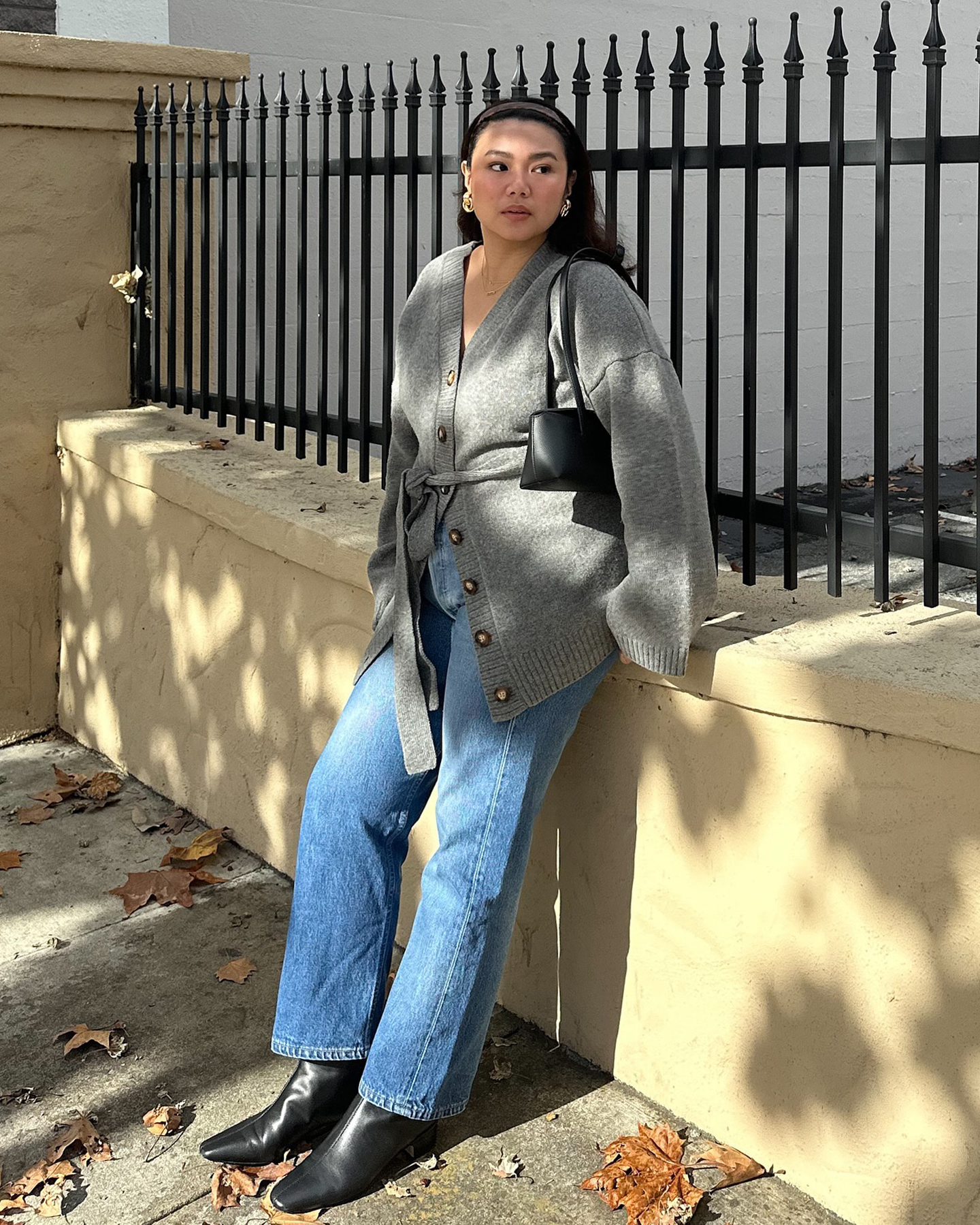 fashion influencer Marina Torres poses on a sidewalk wearing a headband, chunky two-tone earrings, Freya black shoulder bag, gray belted cardigan sweater, straight-leg jeans, and black boots