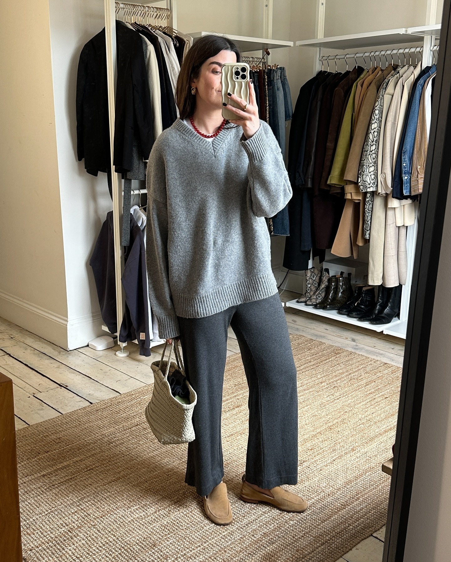 British style influencer Anna Howard of The Anna Edit posing for a mirror selfie wearing a red beaded necklace, a gray oversize v-neck sweater, gray knit lounge pants, an ivory Dragon Diffusion woven tote bag, and tan suede loafers